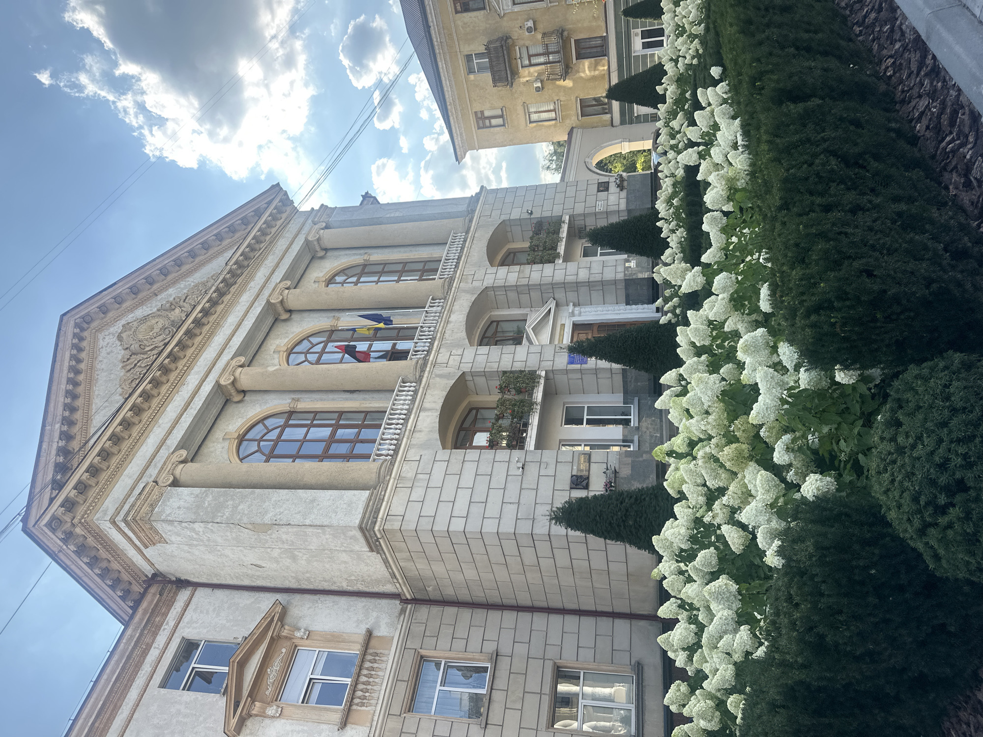 The image shows the Ternopil City Council building, a grand, neoclassical structure located in Ternopil, Ukraine. The building's facade is made of light-colored stone blocks and features tall, arched windows with dark wooden frames. A colonnade with Corinthian columns supports a triangular pediment adorned with ornate carvings. In front of the building, a manicured garden features neatly trimmed hedges and a large bed of white hydrangeas in full bloom. Several conical evergreen trees are strategically placed throughout the garden.
The flags of Ukraine and Ternopil are visible flying from a balcony on the second floor. To the right of the city council building, a neighboring yellow building with balconies and windows can be seen. The sky above is a mix of blue sky and dramatic clouds, with visible power lines crossing overhead, indicating an urban setting. The lighting suggests it is daytime, likely late afternoon, with the sun casting shadows. No people are visible in the image.