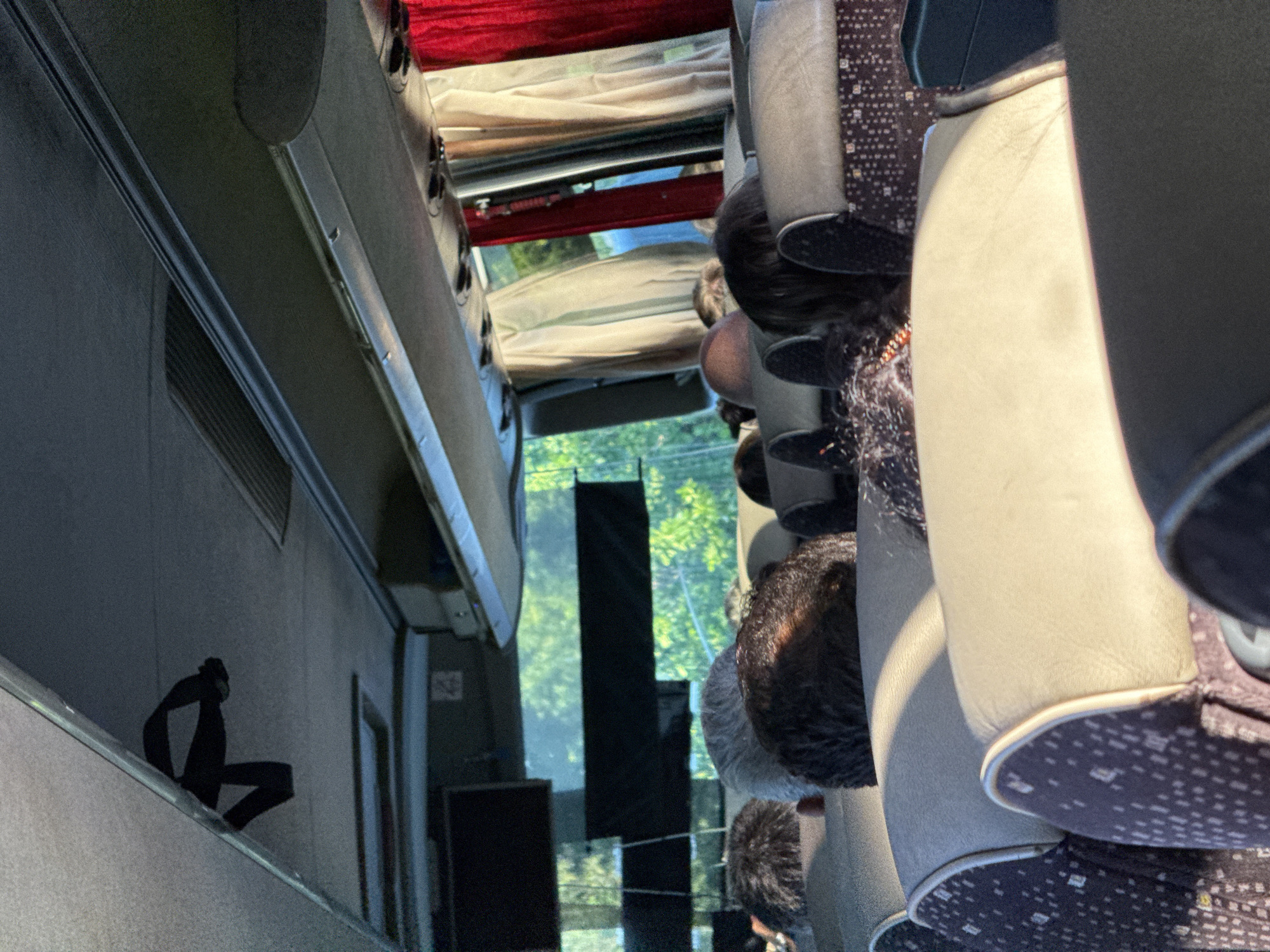 The image shows the interior of a bus, likely in the city of Kiosevtsi, Bulgaria, with passengers seated. The perspective is from a passenger looking forward. Overhead compartments and a strap are visible. Curtains, possibly red and beige, are drawn on the windows. The seats are upholstered, with some showing a dark pattern. The heads and upper bodies of several passengers are visible, their faces obscured by the angle and seating arrangement. Outside the windows, trees and foliage suggest a natural, possibly rural or suburban, environment. The lighting indicates it is daytime, with sunlight casting shadows inside the bus.