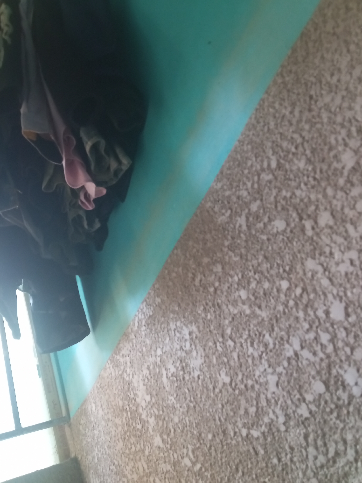 In Jalingo, Nigeria, a laundry area features a turquoise wall juxtaposed with a textured, light brown concrete surface. Clothes, including a pink garment, are hung, partially obscuring a window with metal bars. The lighting suggests an indoor or covered outdoor space.