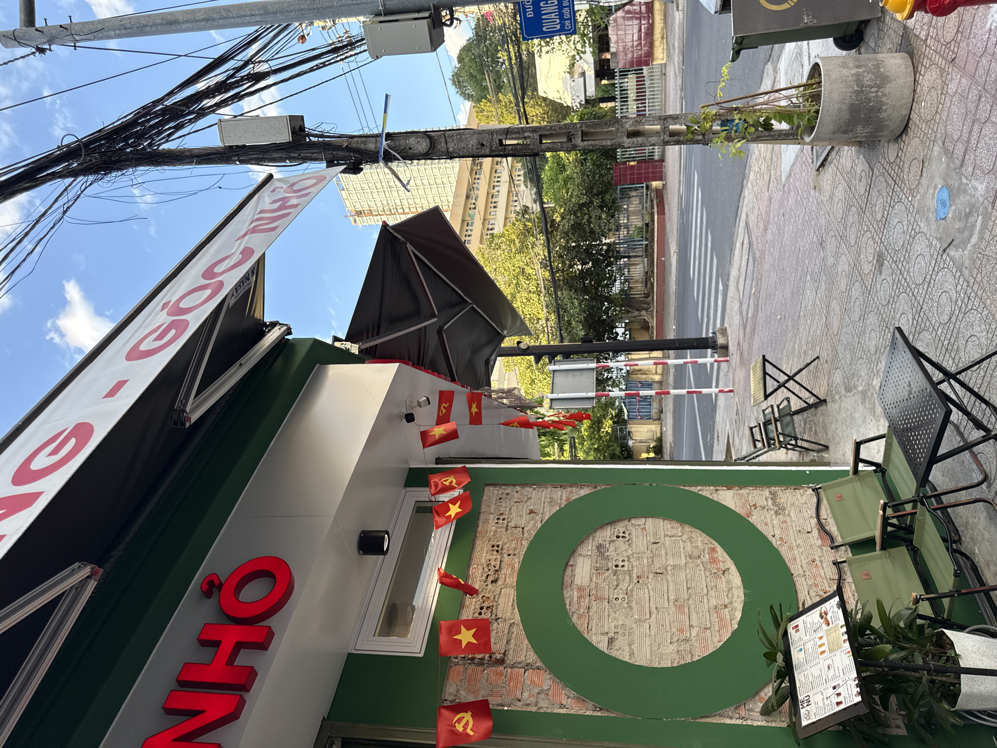 The image shows the exterior of a cafe in Nha Trang, Vietnam. The cafe has a green facade with red lettering that reads "NHỎ". Awnings are extended over the storefront. Small red flags with yellow stars and hammers and sickles hang on a string along the side of the building. A large green circle is painted on a section of exposed brick wall. Two green camping chairs and a small black table are set up on the sidewalk in front of the cafe, next to a menu stand with a potted plant. Across the street, there is a pedestrian crossing, a sidewalk with a large concrete planter containing a thin vine, and a utility pole covered in electrical wires. In the background, there are trees and a tall, light-colored building. A blue street sign with white lettering is visible on the utility pole. The sky is blue with scattered clouds, and the sun appears to be setting or rising, casting long shadows.