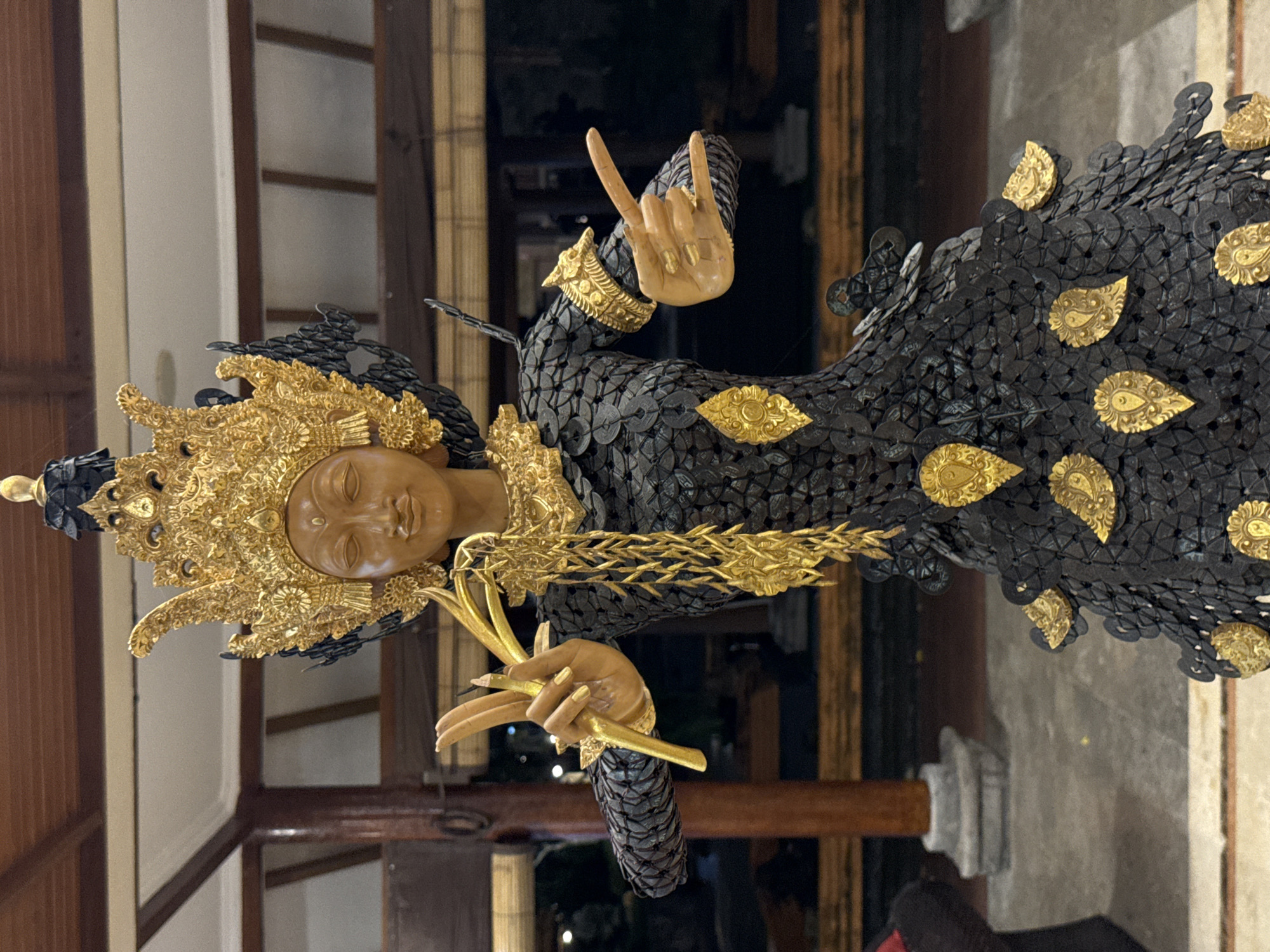 This is a medium shot, eye-level, full-body photograph of a statue resembling a Balinese dancer. The statue is made of dark, overlapping, circular elements, possibly coins, with gold accents. It wears an ornate, golden headdress and elaborate gold jewelry, including a necklace and armbands. The statue's hands are in graceful, expressive poses. It is positioned in what appears to be a covered outdoor or semi-outdoor area with wooden beams and railings visible in the background. The flooring is tiled. The statue is likely located in Kuta Selatan, Indonesia, given the context of Balinese artistry.