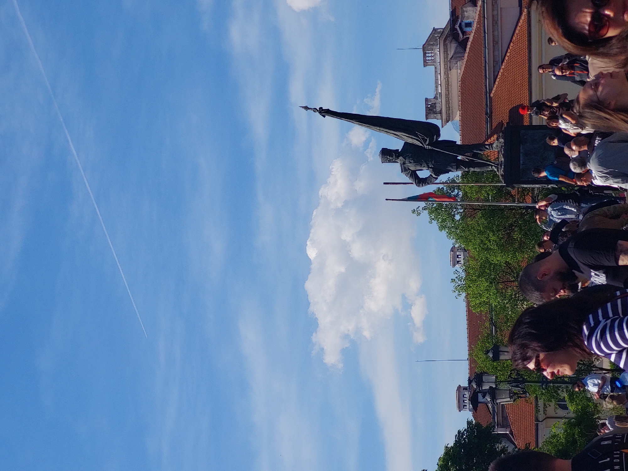 The image shows a daytime scene with a blue sky featuring a contrail and scattered clouds. A statue of a soldier holding a flag stands prominently in the midground, with the flag itself being dark in color. Behind the statue are buildings with red tiled roofs and greenery, suggesting an urban environment. A crowd of people is visible on the right side of the image, with faces and partial bodies indicating they are observing the statue. Flags, possibly national flags, are positioned near the statue. The location is Unknown, Unknown.