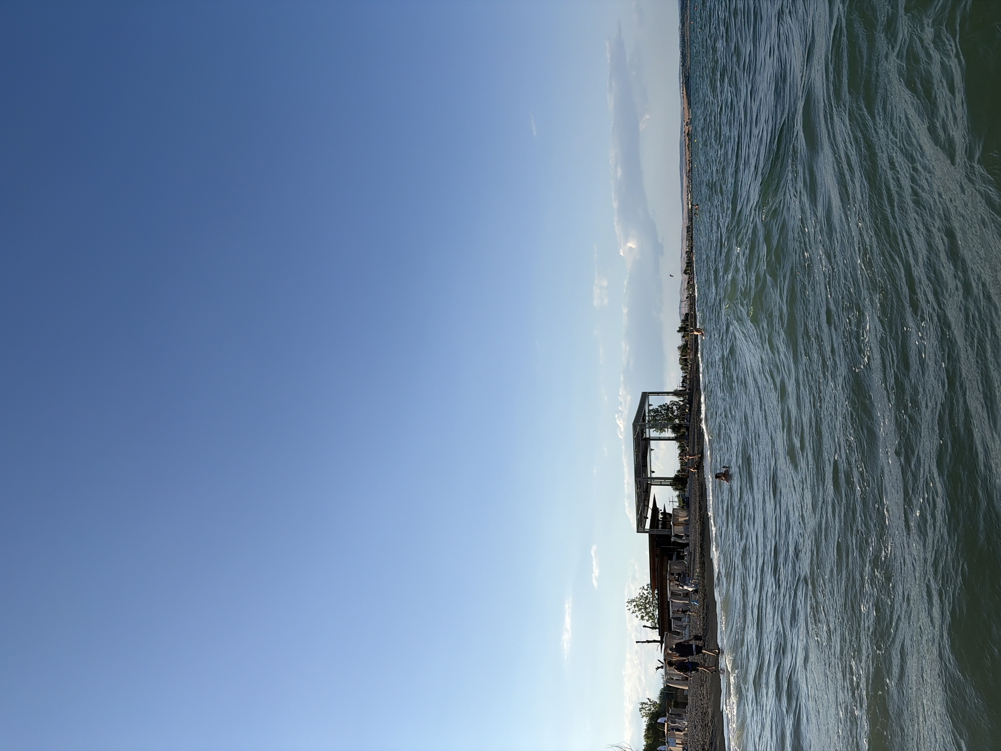This is a vertical, eye-level shot of a beach scene, likely in Burgas, Bulgaria, with the sea in the foreground and a populated coastline in the background. The sky is a clear, deep blue, with a few wispy white clouds scattered near the horizon. The sea is choppy, with small waves creating white foam.
In the foreground, the dark blue-green water fills the lower half of the frame. A person is swimming in the water on the right side of the frame.
The middle ground features a beach made of dark pebbles, with several people walking and standing along the shore. A modern, open-sided structure with a flat roof and metal supports is visible on the beach, with several white canopy-covered seating areas in front of it. Trees are also present along the coastline.
The background shows a distant shoreline with buildings and vegetation, receding into the haze under the bright sky. The overall impression is a lively summer day at the beach.