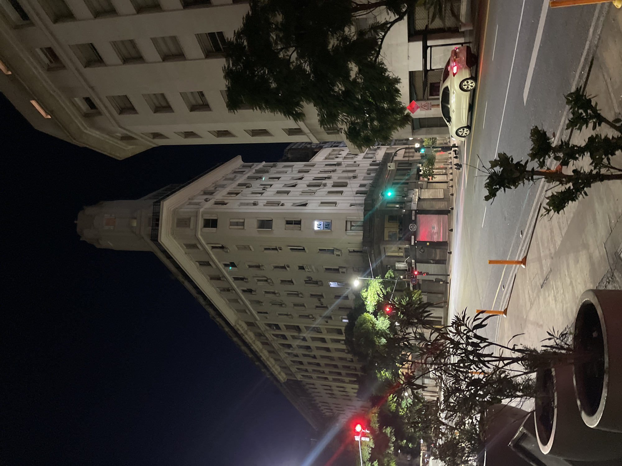 At night, the image captures a street scene in Buenos Aires, Argentina. Several multi-story buildings dominate the view, with numerous windows visible. The sky transitions from dark to bright, possibly due to the presence of city lights. A street with traffic cones is partially visible, and a vehicle is parked along the street. Planters with foliage are positioned on the sidewalk, and trees are also present. Traffic lights are seen in the scene.