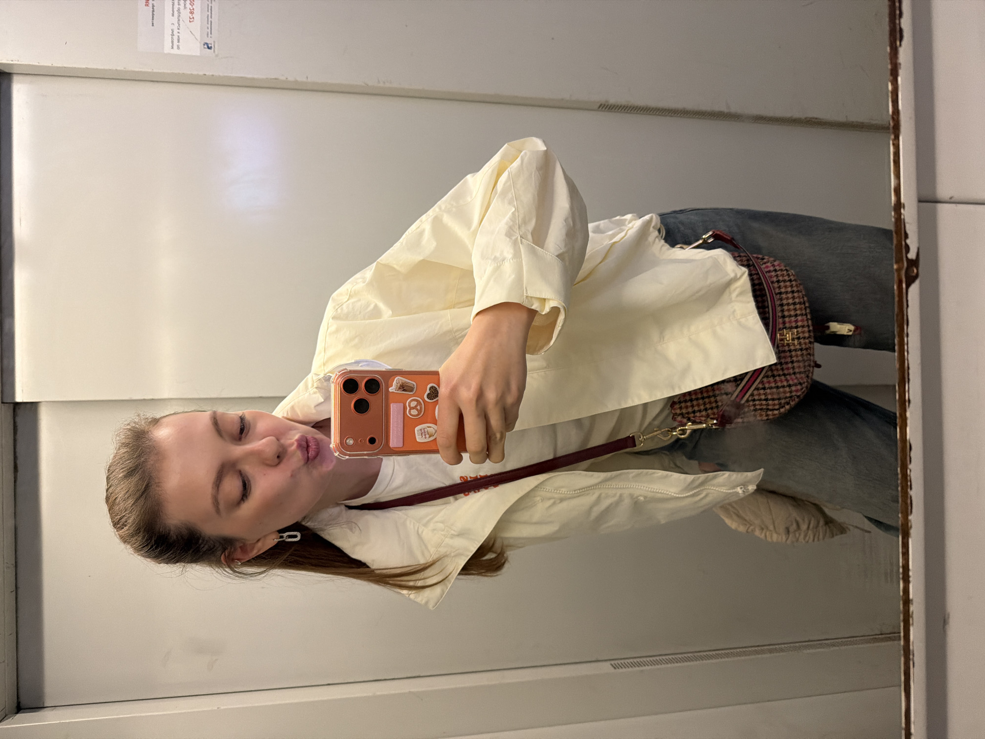 A young woman is taking a selfie in a mirror. She has light brown hair, light skin, and is wearing a white shirt and jeans. She is holding a cell phone with a brown case. A brown bag hangs over her shoulder. The background appears to be a white wall, possibly inside an elevator. A small sign is visible in the upper left corner of the frame. The location is Khimki, Russia, based on the signage.