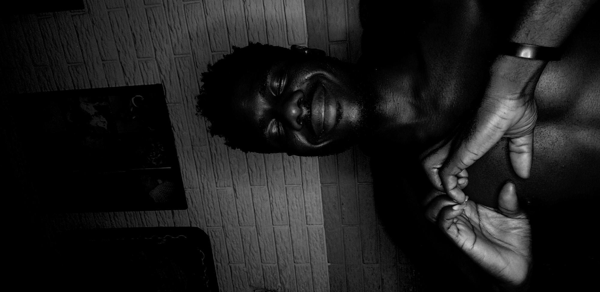 A high-contrast, black and white photograph captures a person, likely male, from the chest up, positioned diagonally across the frame. The subject has dark skin and short, curly hair, and is shown with a joyful expression, smiling or laughing, with eyes closed or squinting. Strong lighting illuminates parts of the face, including the forehead, nose, and lips, creating a luminous effect, while other areas of the face and upper body recede into deep shadows.
In the foreground and to the right, two hands are gently interlocked or touching, suggesting affection or connection. One wrist is adorned with a simple, dark-toned bracelet. The hands are also brightly lit, highlighting their texture and form.
The background features a textured wall, possibly brick or patterned wallpaper, visible in varying shades of grey on the left and above the subject's head. Towards the far left, indistinct dark shapes are visible on what appears to be a shelf or surface, largely obscured by shadow. The dramatic lighting and monochrome palette emphasize the subject's emotional expression and the intimate gesture of the hands. No text is visible in the image.