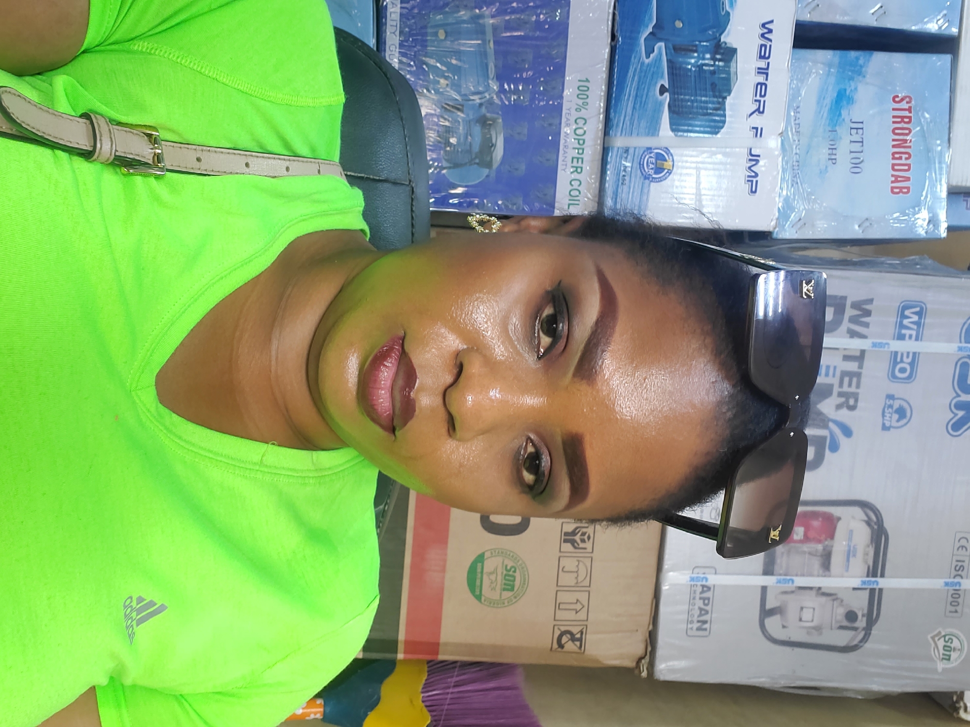 A medium shot depicts a dark-skinned woman wearing a neon green Adidas t-shirt, tan belt, and black Louis Vuitton sunglasses. She is wearing dark lipstick and eyeshadow. She is positioned in front of a stack of boxes. The text on the boxes reads "STRONGDAB, JET100, 1.0HP" and "WATER P JMP". Another box reads "100% COPPER COIL". A box also has the text "WATER WF 20, 5.5HP". The logo for Standards Organisation of Nigeria (SON) appears on some of the boxes. A partial view of what may be a purple broom is visible on the bottom left of the frame. The background suggests an indoor commercial space, potentially in Lagos, Nigeria, given the SON logo on the boxes.