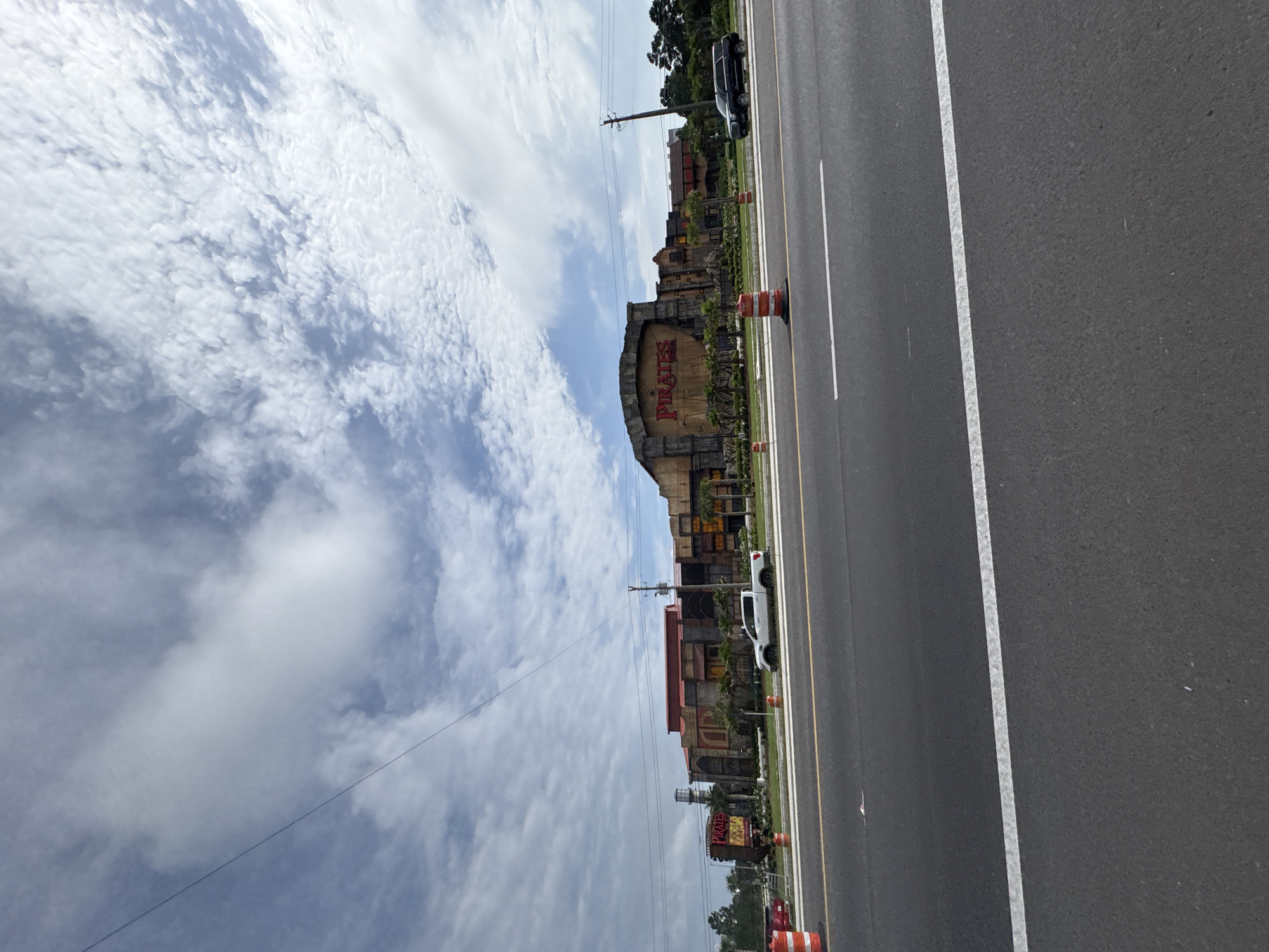The image shows a view of the "Pirates Island Adventure Golf" in Panama City Beach, Florida, USA, from the side of a road. The building features themed architecture with the word "PIRATES" prominently displayed. A multi-lane asphalt road with yellow and white lane markings runs parallel to the attraction. A few vehicles are present on the road. Above, a cloudy sky fills the upper portion of the frame. Construction cones are positioned along the edge of the road near the building. Power lines stretch across the sky above the attraction. The environment appears to be a sunny day.