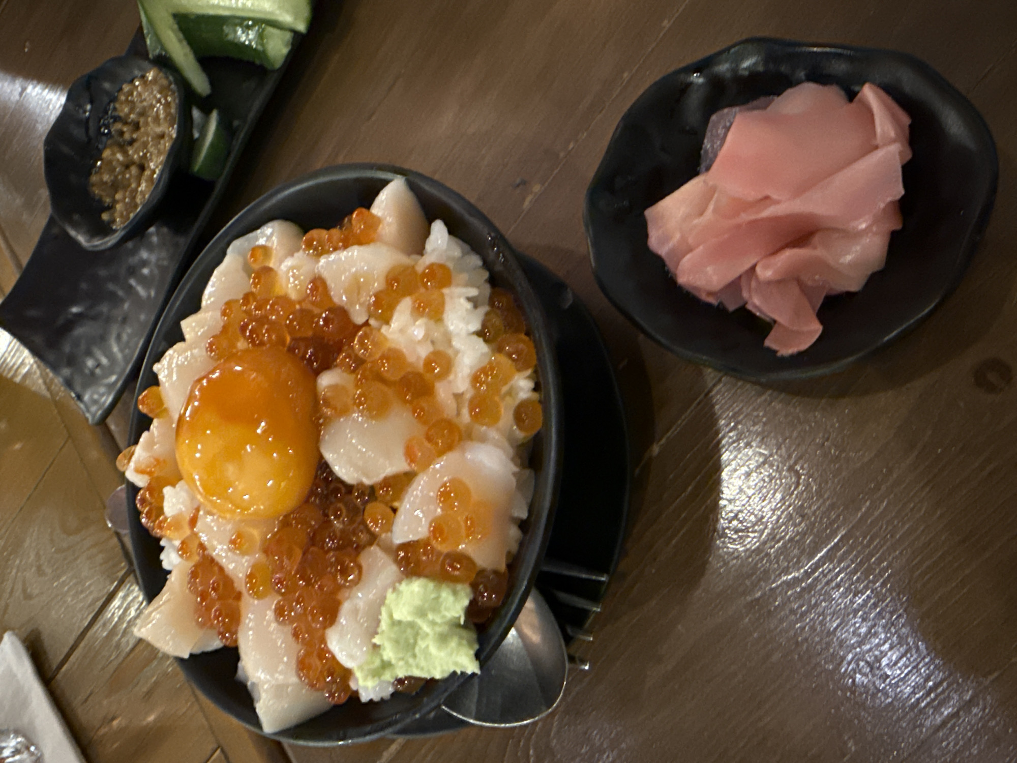 The image shows a meal on a wooden table, likely in a restaurant. There is a black bowl containing rice, topped with fish eggs (likely salmon roe), scallop sashimi, a raw egg yolk, and a dollop of wasabi. To the side, in another black dish, is pickled ginger. A long black plate holds a small dish of a brown sauce, and several cucumber slices. A spoon is next to the rice bowl. The setting could be a Japanese restaurant in Bangkok, Thailand, though there are no distinctive architectural elements to confirm the city. The scene appears to be a close-up of a table setting.