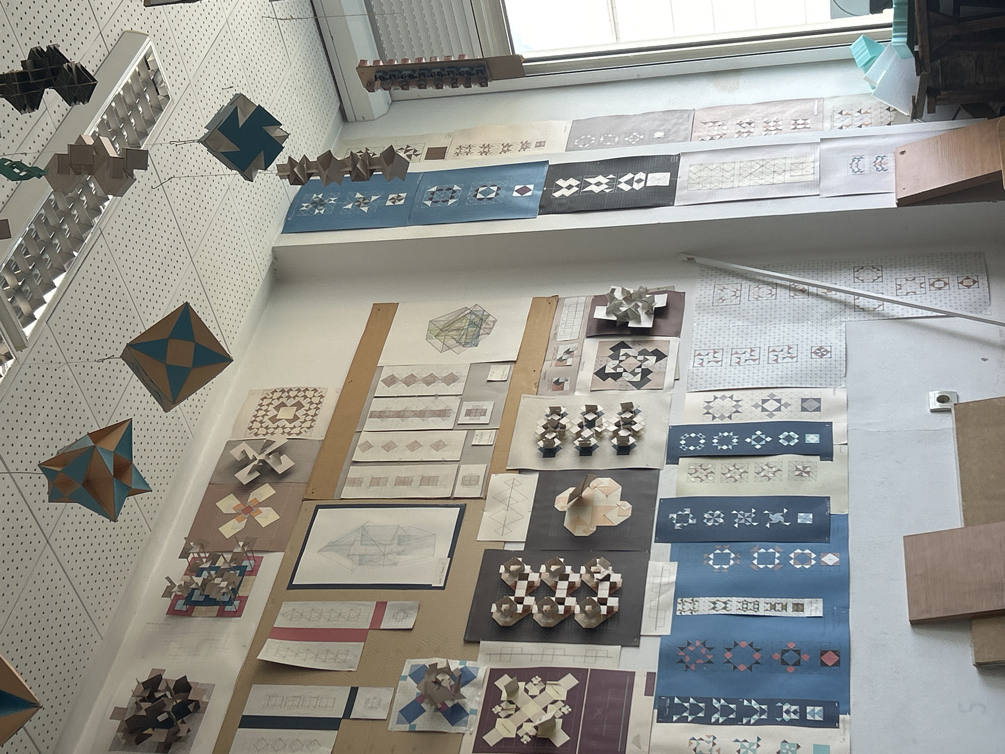 This image captures an interior scene, likely a design studio, architecture workshop, or art classroom, showcasing an exhibition or display of geometric and modular design projects. No people are visible.
The setting features white walls, a perforated acoustic ceiling with fluorescent light fixtures, and a large window at the top providing natural light. Various geometric paper models are suspended from the ceiling, resembling polyhedra or intricate modular forms, some in teal and brown, others in neutral cardboard tones. More such models, often composed of repeated units, are arranged on large display boards or tables against the wall.
The walls and display surfaces are covered with numerous sheets of paper featuring detailed architectural drawings, geometric patterns, and tessellations. These patterns vary in complexity and color, often utilizing shades of blue, brown, grey, and white, depicting arrangements of squares, stars, diamonds, and triangular elements. One visible drawing shows a multi-perspective architectural sketch. Other papers display sequences of patterns or studies of structural forms. Small wooden blocks are also present on some surfaces, possibly as part of a model or display element.
The arrangement suggests a curated exhibition or an academic presentation of design work. The bright lighting indicates it is daytime. The location context is Sofia, Bulgaria, implying this space is situated there. No discernible text is clearly readable on the documents due to the angle and resolution, although some papers appear to have annotations or titles.