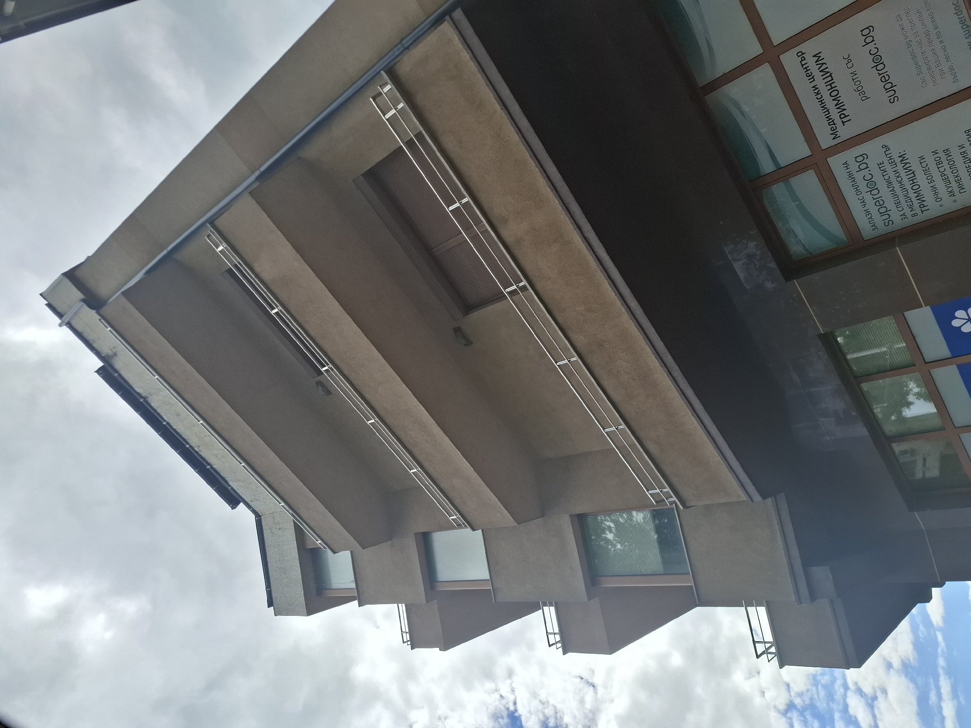 The image is an exterior, low-angle, wide shot of a modern, multi-story building with a brown facade and multiple balconies. The building features horizontal metal railings on each balcony. The sky is overcast with some patches of blue sky and clouds visible. On the ground floor of the building, a window displays signage for "Медицински център ТРИМОНЦИУМ" (Medical Center TRImONTSiUM) and advertises online booking via "superdoc.bg". The building appears to be located in an urban environment, although no specific city details are visible.