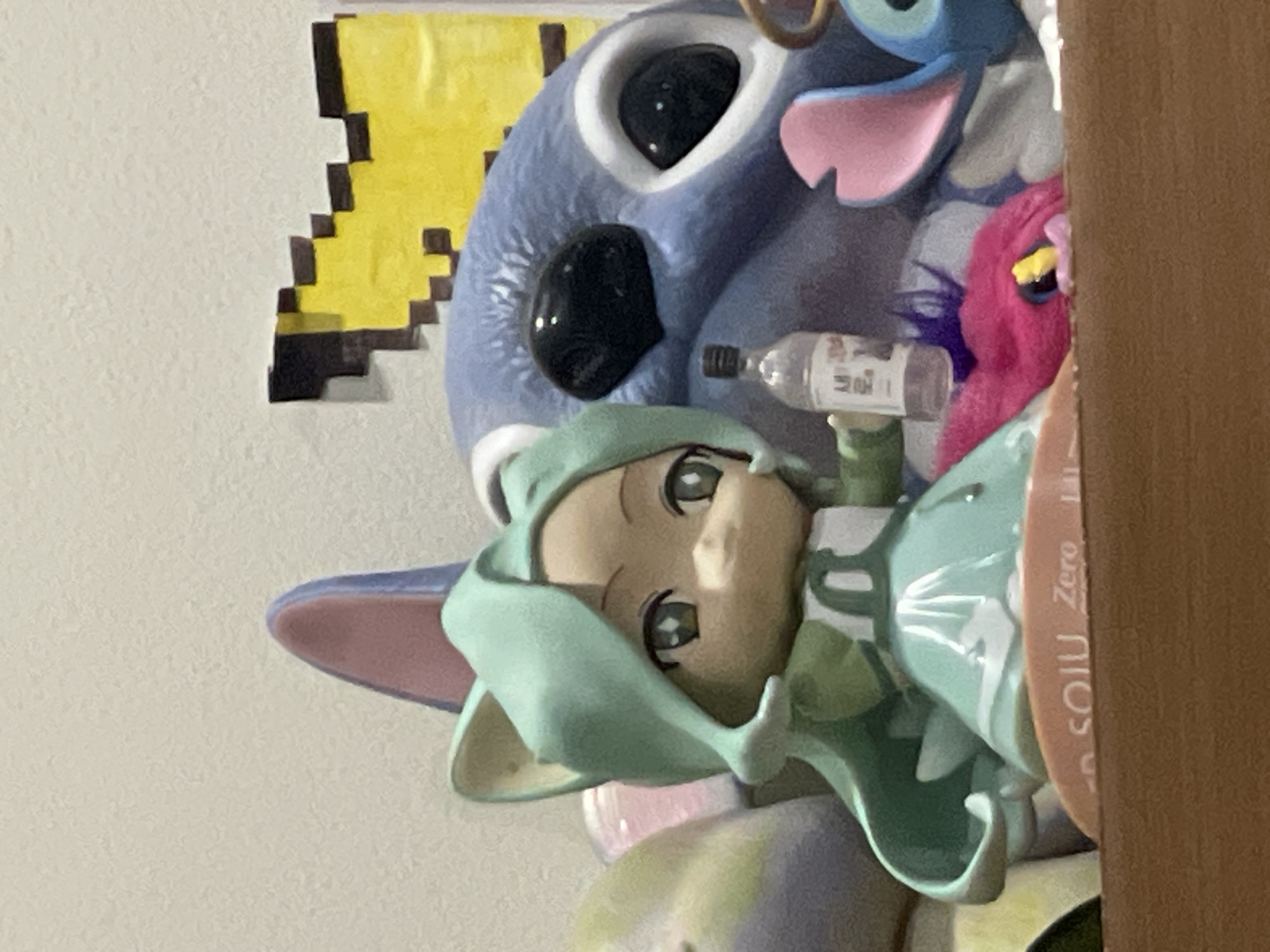 The image shows a close-up of several collectible figurines displayed on a wooden shelf. In the foreground, a small anime-style figurine with green hair and cat ears is holding a miniature bottle. Behind it, a larger blue character, resembling Stitch from Disney's Lilo & Stitch, is visible. To the left and right of these main figures, other colorful figurines and plush toys are partially visible. The background appears to be a textured wall with pixelated yellow and black graphic art. The lighting is soft and likely from an indoor source. There is no indication of a specific location in Denpasar, Indonesia, or any context beyond the arrangement of the displayed items.