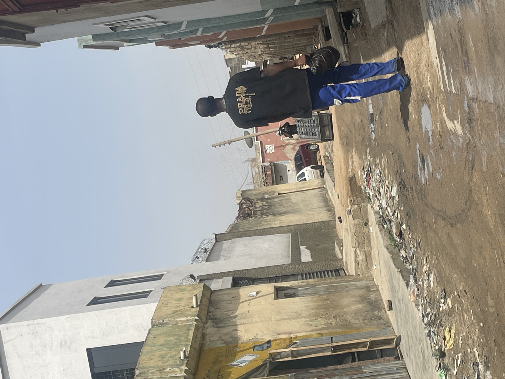 The photo shows a man in blue pants and a black shirt that says "PRADA" on it, walking down a street with buildings and garbage along the side of the road. The buildings are various shades of white, gray, and yellow. There are a few cars visible in the distance. The location appears to be a city.