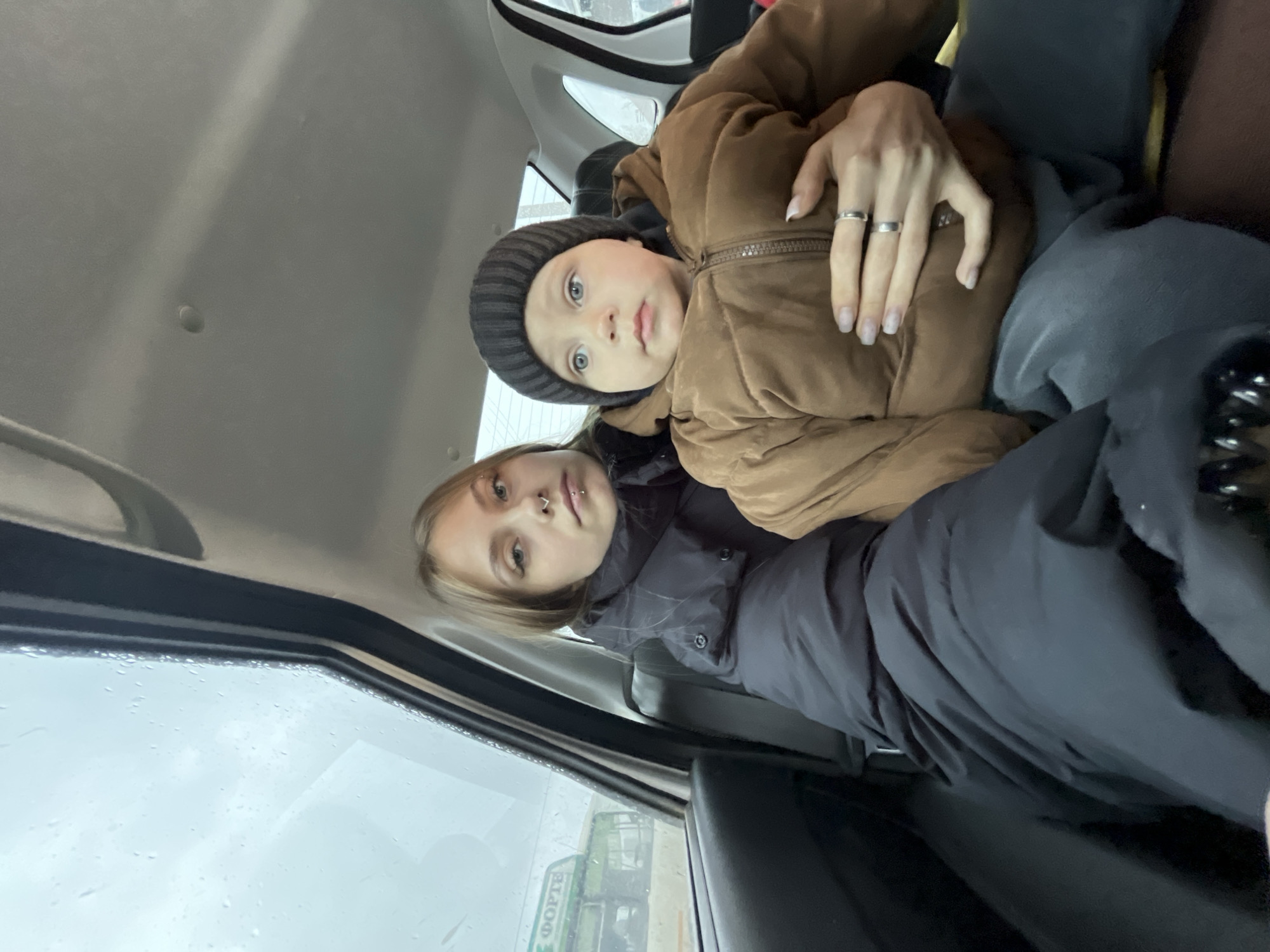 This image features a young woman and a small child seated inside a vehicle, both looking directly towards the camera. The woman, positioned closer to the viewer, has light-colored hair, a nose piercing (septum), and light-colored eyes. She wears a dark puffer jacket and has two silver-toned rings on her left hand's ring and middle fingers, with light nail polish visible. She appears to be holding or embracing the child.
The child, likely a young boy, wears a brown puffer jacket and a dark striped beanie, also possessing light-colored eyes. Both individuals display neutral expressions.
The setting is the interior of a car or van, evident from the headliner, window frame, and seat contours. A side window on the left reveals an exterior view. The weather outside is overcast, and numerous water droplets on the window suggest it is raining or has recently rained. Through the blurry glass, an urban or suburban landscape is visible.
A distinct detail is the partial view of Cyrillic text on a building or vehicle outside the window, which reads "ФОРТЕ" (FORTE). This detail, combined with the provided context, places the scene in Фоки, Russia, during daytime under grey, possibly rainy, conditions.