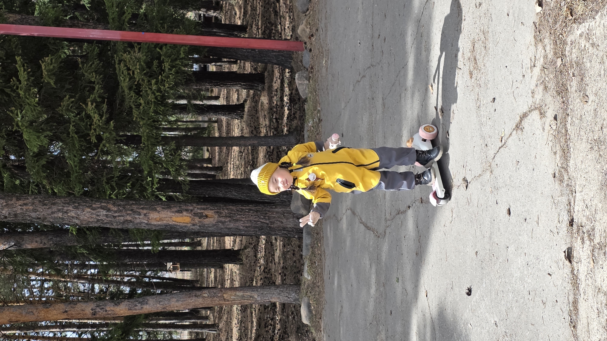 A child in a yellow jacket and hat is standing on a skateboard on an asphalt path. The child is holding a small pink object in their hand. The path is bordered by trees on one side and a gravelly area on the other. In the background, there are trees and a red structure. The location may be in Lensk, Russia.