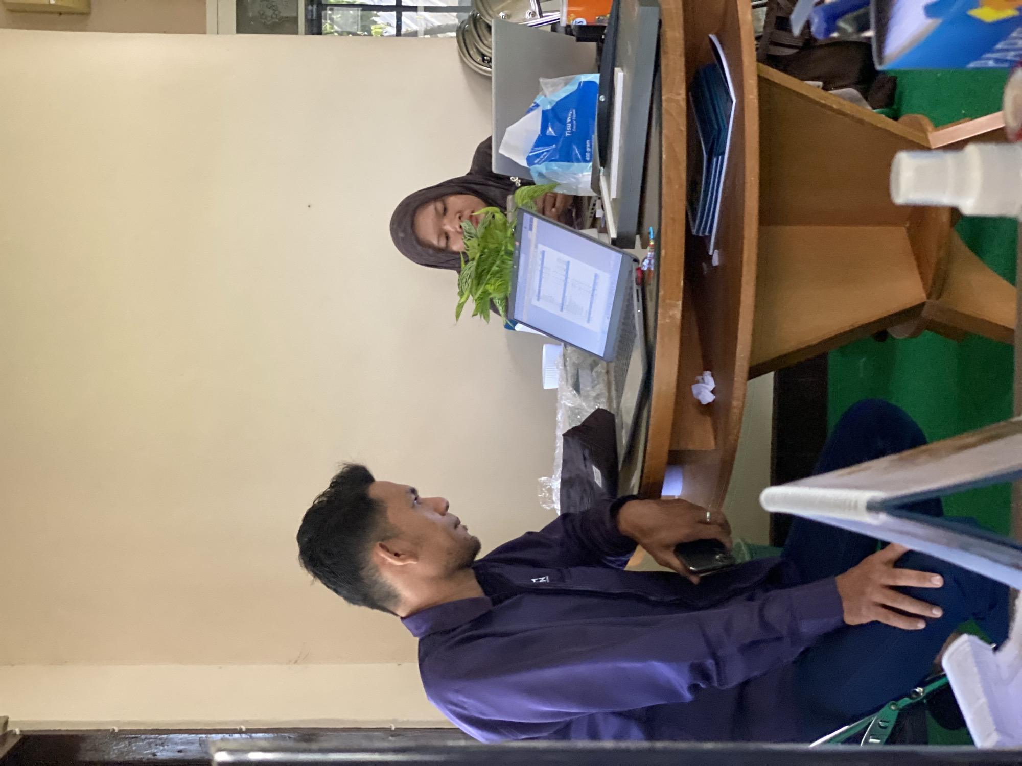 The image depicts an indoor scene, likely an office or meeting room, situated in Banjarbaru, Indonesia. A man in a purple collared shirt and dark pants is seated and looking at a laptop on a round wooden table. He is holding a dark smartphone in his left hand. Opposite him, a woman wearing a dark hijab and a brown top is also seated, looking down at her lap while holding a pen. She is positioned behind the laptop.
On the table, several objects are visible: two laptops, a blue package of tissues, a potted green plant, a paper cutter, and various papers and electronic devices. The table has a distinctive, geometrically shaped wooden base. In the foreground, a white foldable stand is partially visible. The background is a neutral-colored wall. The floor is covered in green carpeting. The overall lighting suggests an ambient indoor setting, possibly with natural light from a nearby window or opening, hinted at by the light source from the right side of the image.