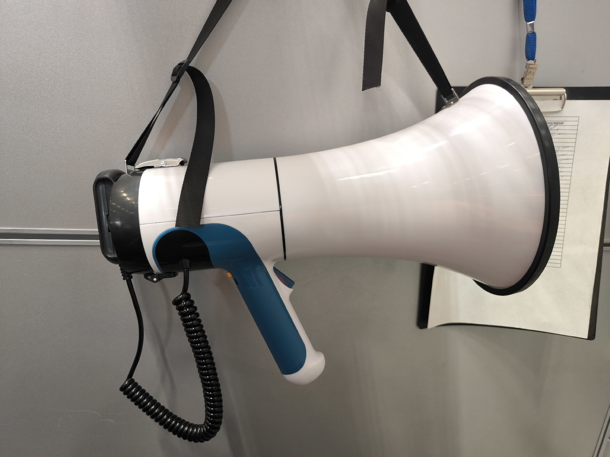 A white and blue megaphone is hanging from a black strap against a gray wall. The megaphone has a coiled black cord attached to its handle. To the right of the megaphone, a clipboard with a document is visible, held by a blue lanyard with a silver clip. The image appears to be an indoor setting.