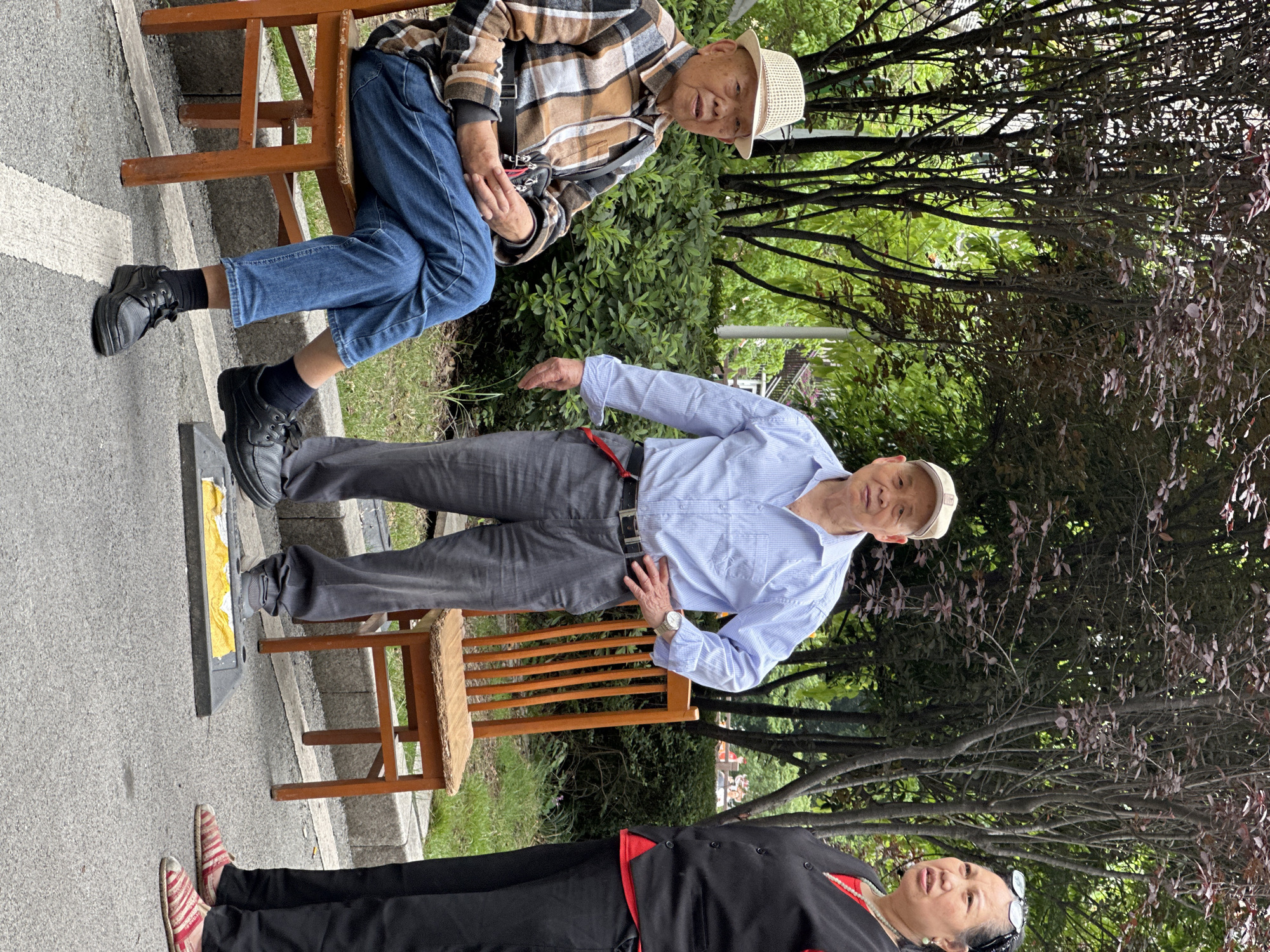 The image shows three people outdoors in Yuzhong District, China.
The top individual sits on a wooden chair, wearing a plaid shirt, blue jeans, black socks, and black shoes. He also wears a straw hat.
In the middle, a second individual stands, wearing a light blue button-down shirt, gray pants, black shoes, and a baseball cap.
The third person lies on the ground, wearing red and white shoes, black pants, and a black top with a red lining.
They are on a paved surface near some trees and bushes.