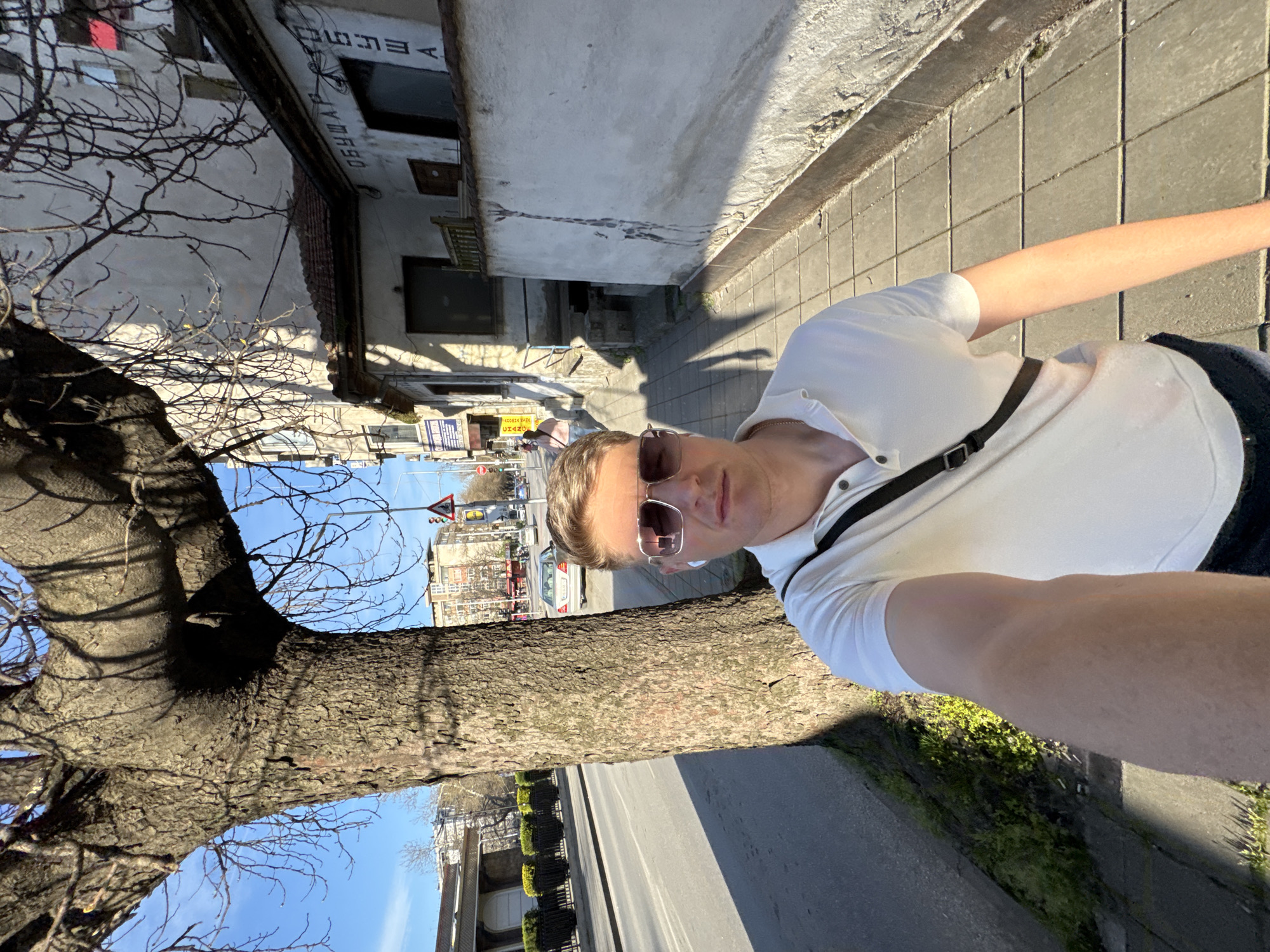 A man in sunglasses and a white polo shirt takes a selfie on a sunny day in Plovdiv, Bulgaria. He is standing on a sidewalk with a tree to his left. Behind him, a street with cars and buildings is visible, including a traffic light. Some buildings show signage. The sidewalk is paved.