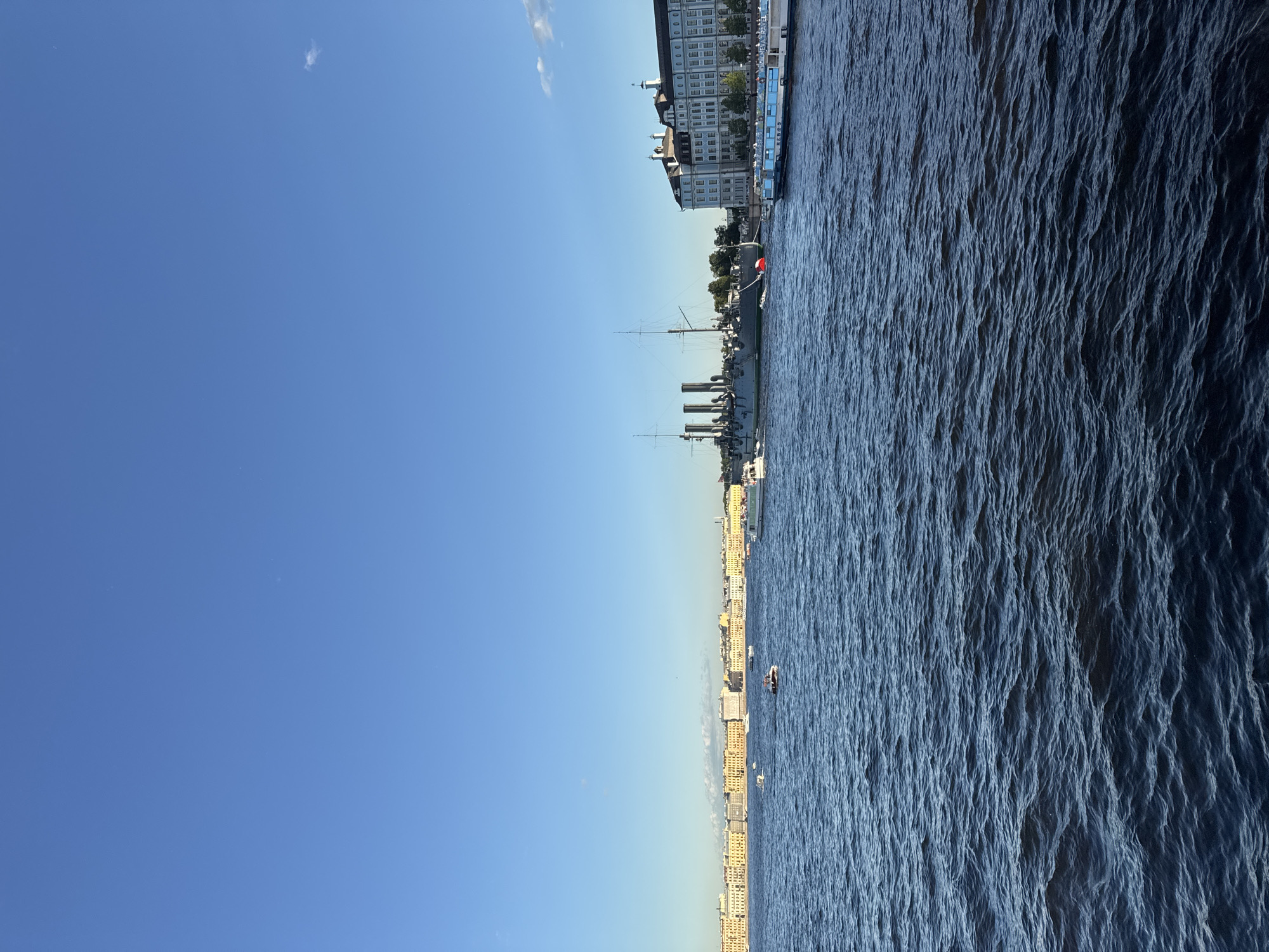 The image depicts a wide, calm body of dark blue water in the foreground, with gentle ripples. In the mid-ground, a large, gray, historical warship with multiple smokestacks and masts is docked. To the right of the warship, a long, white and blue tour boat with people visible on its deck is also docked. Beyond the docked vessels, a row of light yellow buildings with arched windows stretches along the distant shoreline. Several smaller boats are scattered across the water, some with people on board. The sky above is a clear, bright blue with a few faint white clouds near the horizon. The overall scene suggests a sunny day in a city with a significant waterfront.