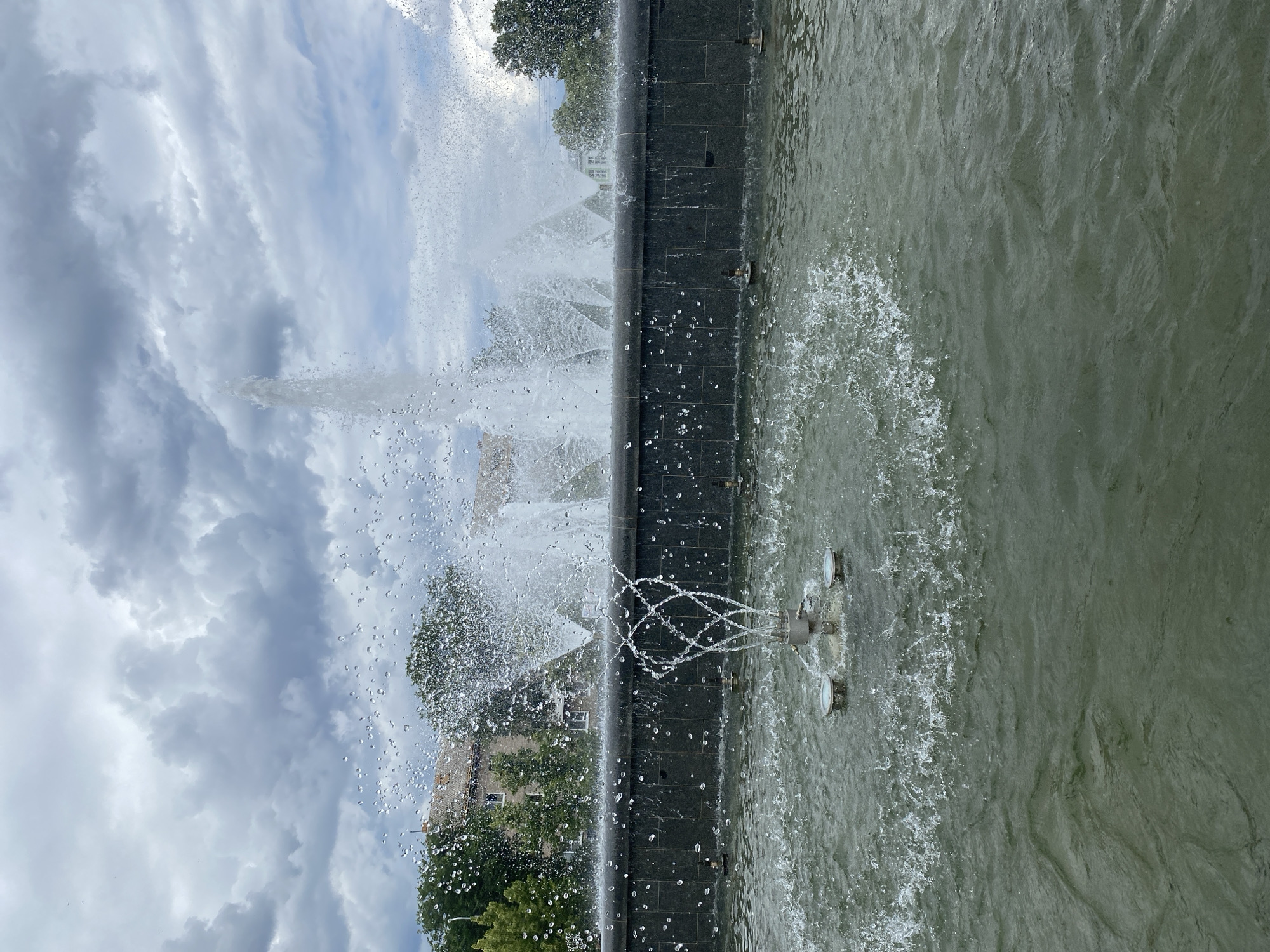 This media file is a photograph of a fountain in Chaikovsky, Russia. The fountain is made of dark stone and features multiple jets of water spraying into the air. The water in the fountain is a murky green color. In the background, there are trees and buildings, as well as a cloudy sky. The photograph is taken from a low angle, looking up at the fountain. The overall mood of the image is peaceful and serene, with the sound of the water being the only thing that could be heard. The time of day is likely afternoon, given the lighting conditions. The weather appears to be overcast, with heavy clouds suggesting the possibility of rain.