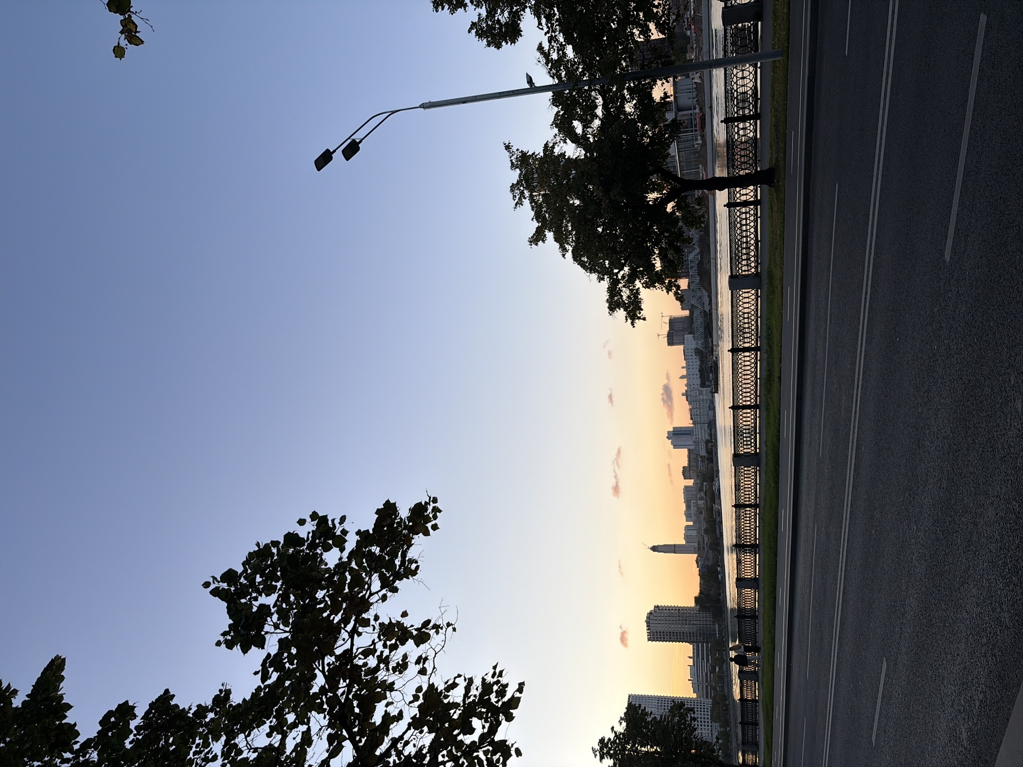 The image depicts a dusk scene along the Moscow River embankment in Moscow, Russia. The sky is a gradient of pale blue to soft orange, indicating the sun has recently set. In the foreground, a two-lane asphalt road with white lane markings stretches across the frame. Adjacent to the road is a grassy strip and a decorative black metal fence with ornate circular patterns, typical of Moscow's embankments.
Beyond the fence, the Moscow River reflects the soft colors of the sky. Two individuals are visible walking along the embankment, silhouetted against the lighter backdrop. Across the river, a cityscape is dominated by a line of modern high-rise buildings. A particularly prominent, tall, slender building with a spire-like structure is centrally located among the skyline. The architecture suggests a contemporary urban environment.
On the right side of the frame, a tall lamppost with two downward-facing lights stands against the sky. Trees with dark foliage are present on both the left and right sides of the image, partially obscuring the view of the sky and buildings. The overall impression is one of a tranquil urban evening.