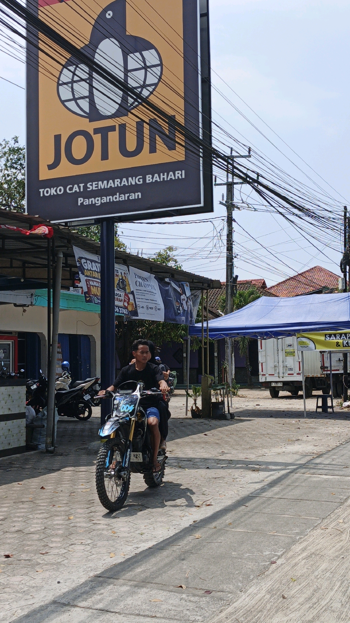 The image depicts a street scene in Desa Wonoharjo, Indonesia, on a sunny day. In the foreground, a young man is riding a dirt bike with blue and black graphics. He is wearing a black t-shirt and blue shorts. The motorcycle has the number "009" on the front. Behind him, other motorcycles are parked. A large sign for "JOTUN" paint is prominently displayed, featuring the brand's logo and text indicating a paint store in Pangandaran. Several banners with promotional messages are hung under a covered area. Further in the background, a white truck is parked under a blue tent, and a yellow and green banner with text is visible. Buildings with red tiled roofs are also part of the environment. The ground is paved with concrete. Utility poles and a network of electrical wires are visible against the sky.