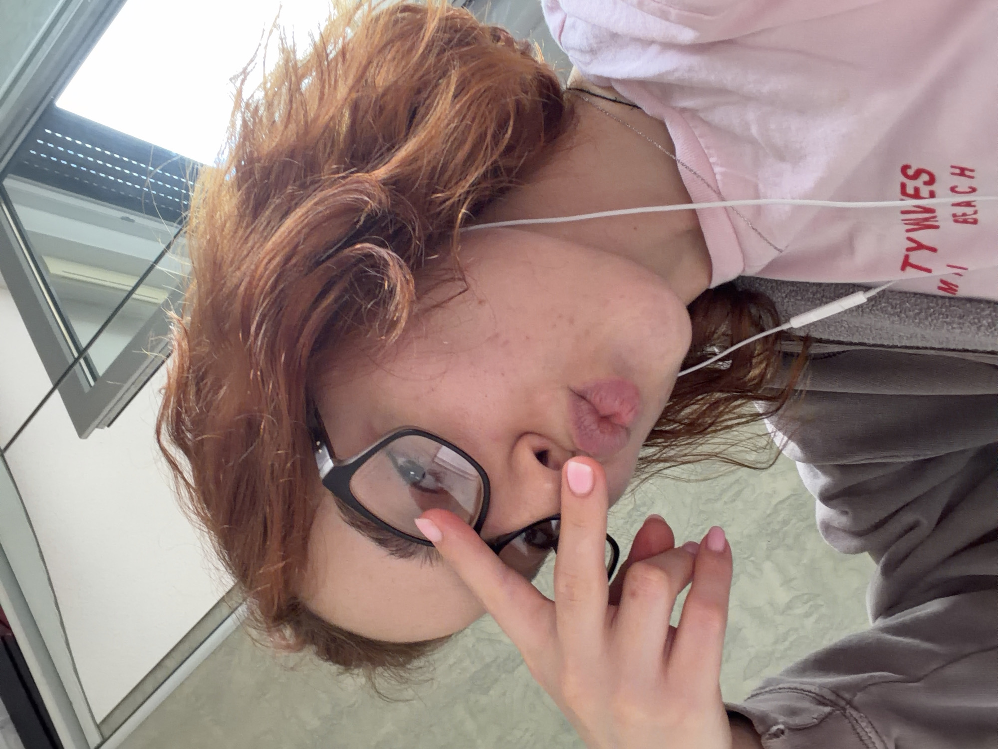 A low-angle selfie captures a young individual, likely female, with reddish-brown, wavy hair and black-framed glasses. The person is making a "kiss face" with pursed lips and has light pink nail polish on their fingers, one of which is raised near their glasses. White wired earbuds are visible, with the cord draped across their neck and a silver chain necklace also present. They are wearing a light pink t-shirt with red text that partially reads "TYWES" and "M I BEACH", layered over a grey hooded sweatshirt or jacket.
The background suggests an indoor setting with modern architectural features. A very bright window or skylight is visible at the top left, indicating daytime with abundant natural light. Below the window are reflective surfaces, possibly glass or mirrors, and light-colored walls or partitions. The image captures a casual and playful moment. The location context provided is Strasbourg, France.