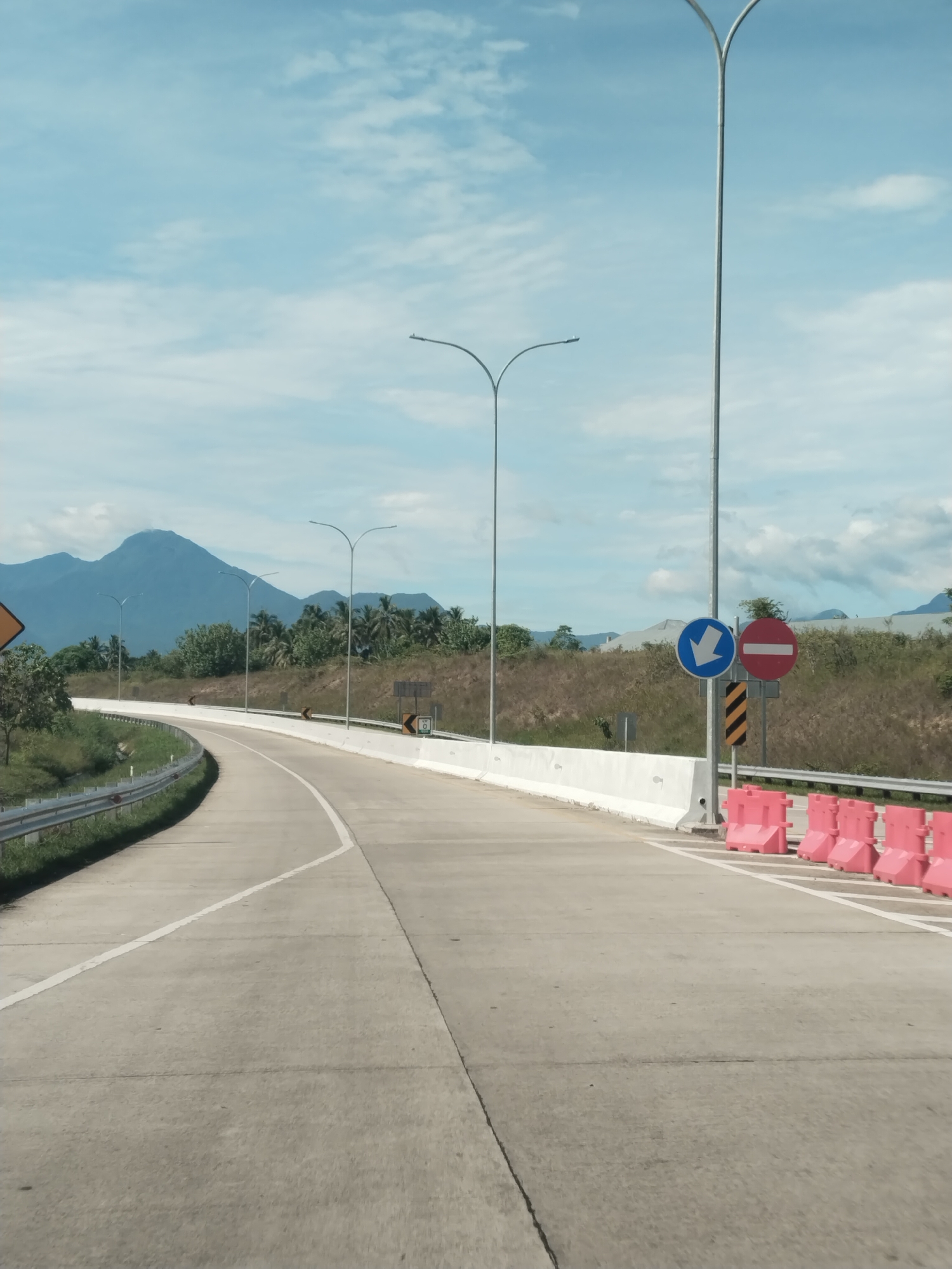 The image shows a paved road in Sihomlhok, Indonesia. The road curves to the left and is marked with white lines. On the right side of the road, there are pink barriers and a signpost with a blue arrow pointing down and a red circle with a horizontal line. Tall streetlights are visible along the road. In the background, there are mountains and trees under a cloudy sky. The scene appears to be taken during the day.