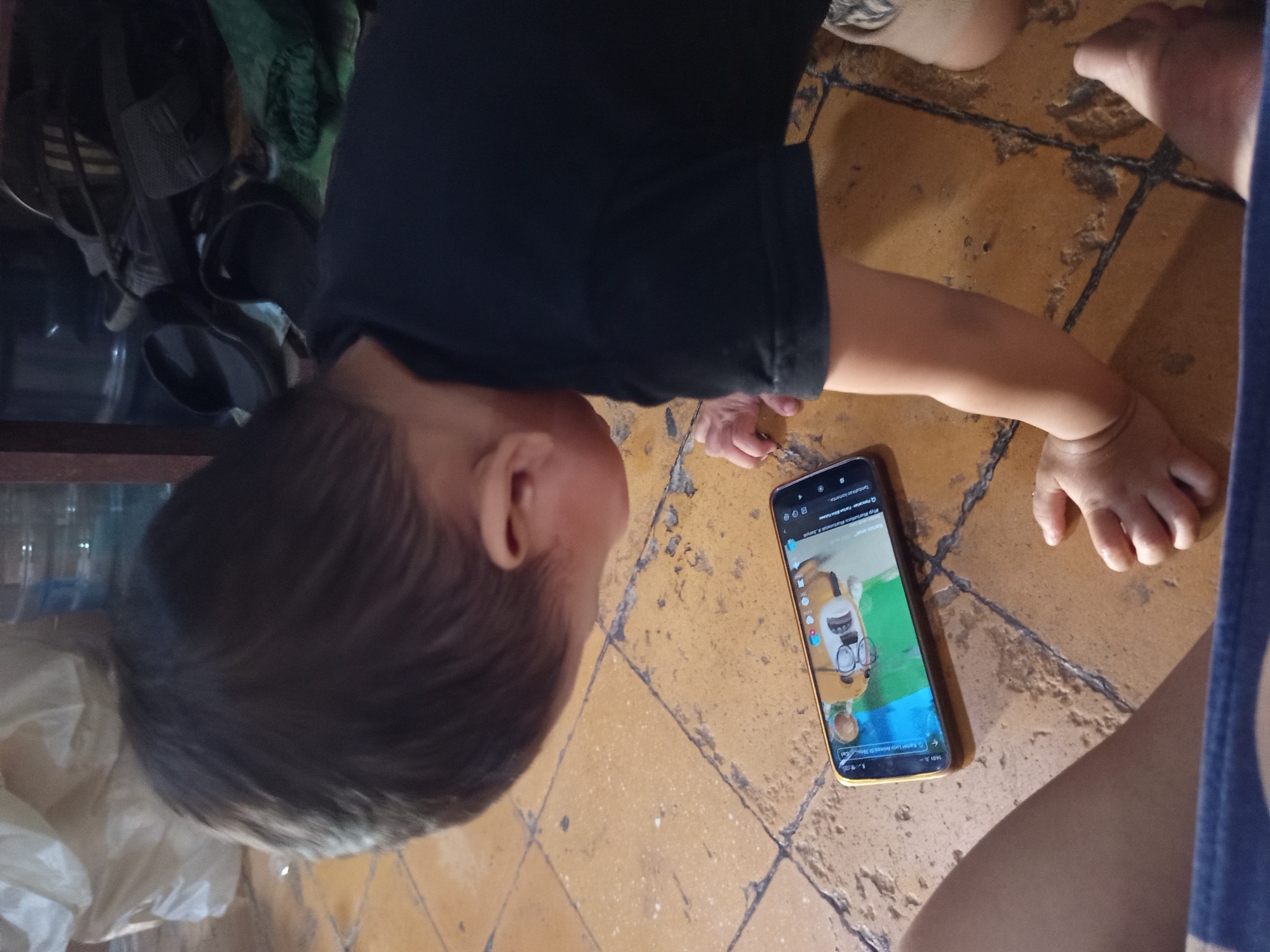 A young child, wearing a black t-shirt, is kneeling on a floor with orange tiles and dark grout. The child is looking down at a smartphone that is placed on the floor in front of them, displaying a game with cartoon characters. The phone has a gold-colored frame. The background shows a blurry environment with what appears to be a large white bag or tarp, and some dark shelves with footwear. The scene is indoors, likely in a home, possibly in the city of Tebel, Indonesia, given the tiles which are common in Indonesian homes. The child's hands are visible, one touching the floor near the phone, and the other reaching out slightly. The lighting suggests an indoor setting with ambient light.