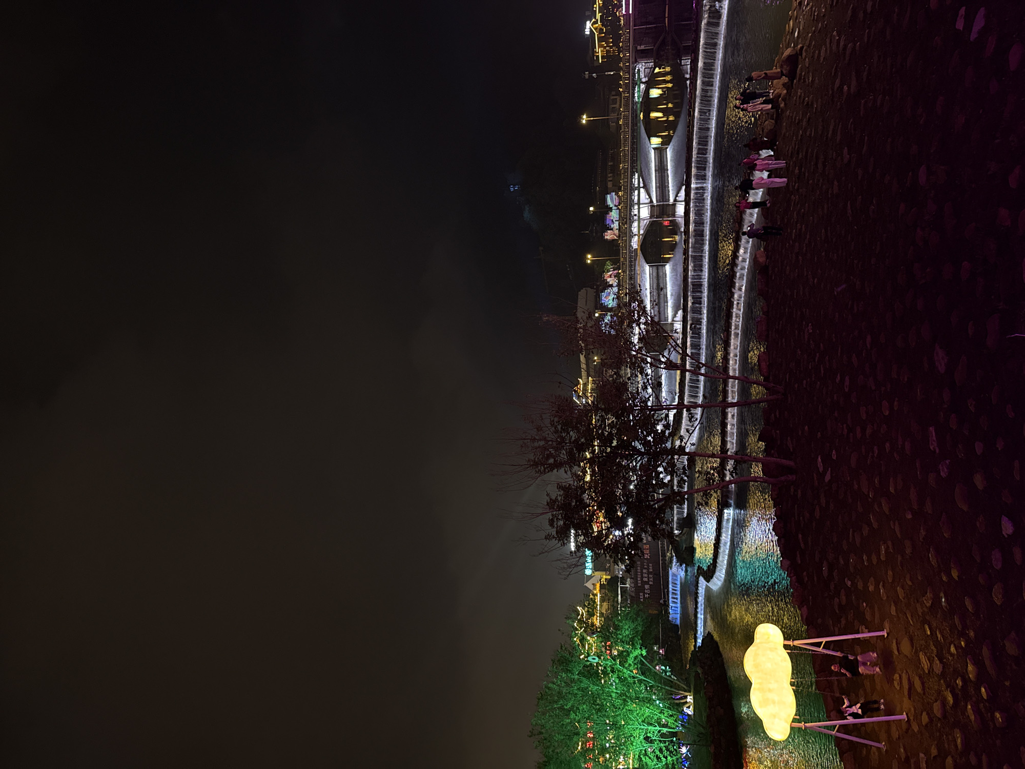 Here's a description of the image:
The image captures a night scene in Wulingyuan, China. A waterfall cascades in the center of the image. Several people stand along the side of the river, observing the scene. The background shows buildings and other structures illuminated with lights. In the foreground, there is foliage, including trees, lit by green lights. A structure resembling an egg is brightly lit. Additionally, there are two people on swings.