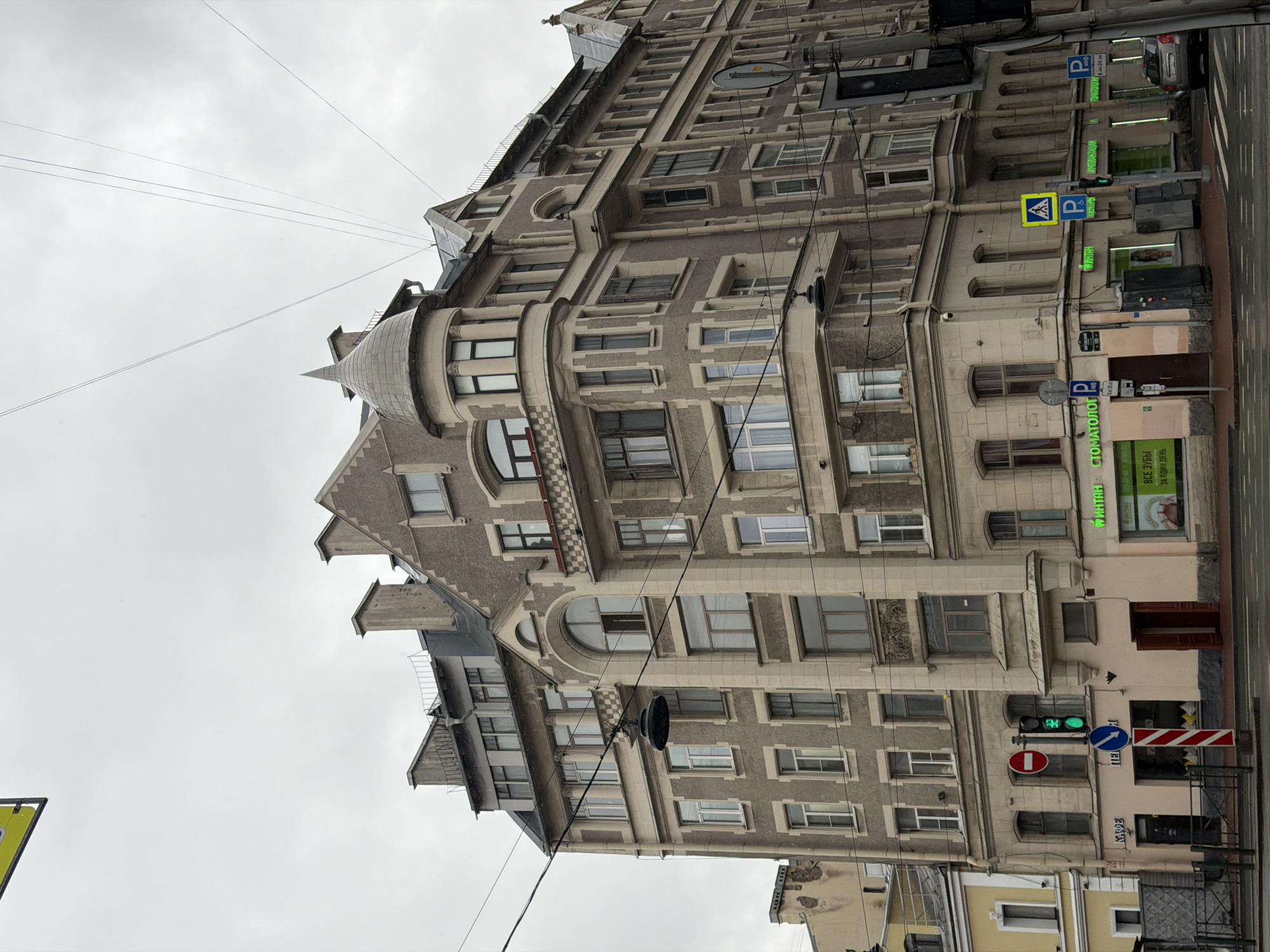 The image depicts a multi-story building, likely an apartment complex, against an overcast sky. The architecture of the building is characterized by varying window shapes and ornate decorations. Signage indicating "Stomatolog" and "Dentist" suggests the presence of dental clinics within the building. Street signs and traffic signals are visible in front of the building. Based on architectural style, signage in Russian, the presence of a pedestrian crossing sign, and other markers, this photo likely depicts a street scene in Saint Petersburg, Russia.