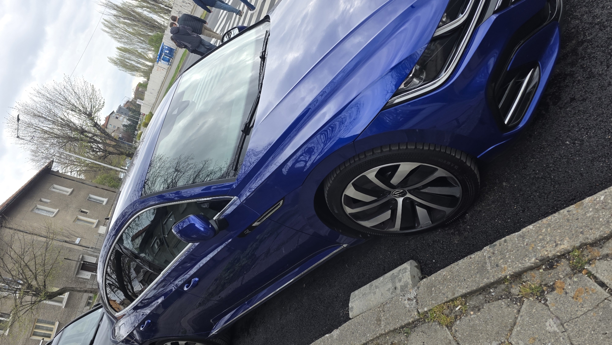 A close-up shot captures a blue Volkswagen car parked alongside a gray concrete curb on an asphalt surface. To the left, the corner of a light-colored building with visible windows is partially seen, with bare trees in front of it. In the background, a glimpse of a street with pedestrians crossing is observed, framed by leafless trees and urban structures. The car's front wheel is prominently displayed, featuring a unique multi-spoke design with the Volkswagen logo at its center. Given the car's license plate containing "E", it's inferred that the photo may be located in Blagoevgrad, Bulgaria.