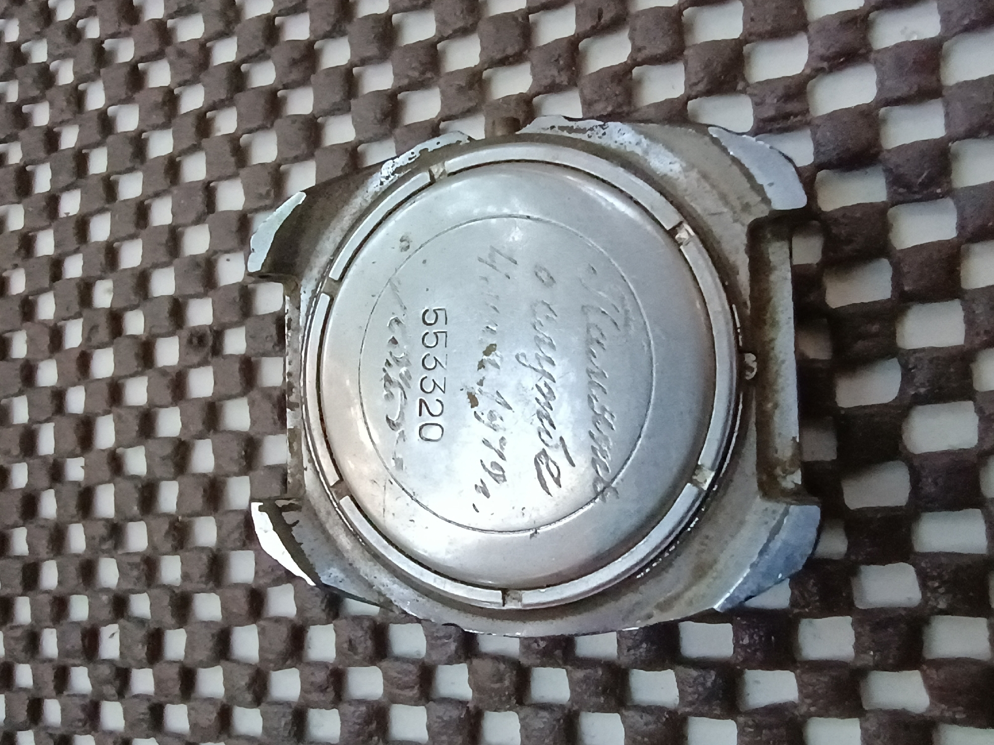 A close-up, overhead shot focuses on the back of a vintage watch case lying on a textured, checkered surface. The watch case is made of silver-toned metal and exhibits signs of wear and age, with some scuff marks and scratches visible on its surface. Engraved on the back of the case are Cyrillic characters and a numerical sequence. The top line of engraving appears to read "553320." Below this, another line of engraving is present, but it is partially obscured and difficult to fully decipher. The checkered surface beneath the watch consists of small, dark brown cubes arranged in a uniform grid pattern. The lighting is overhead, creating subtle shadows and highlighting the textures of both the watch case and the surface it rests upon. The image does not provide any explicit information about the location of Zaporizhzhia, Ukraine.