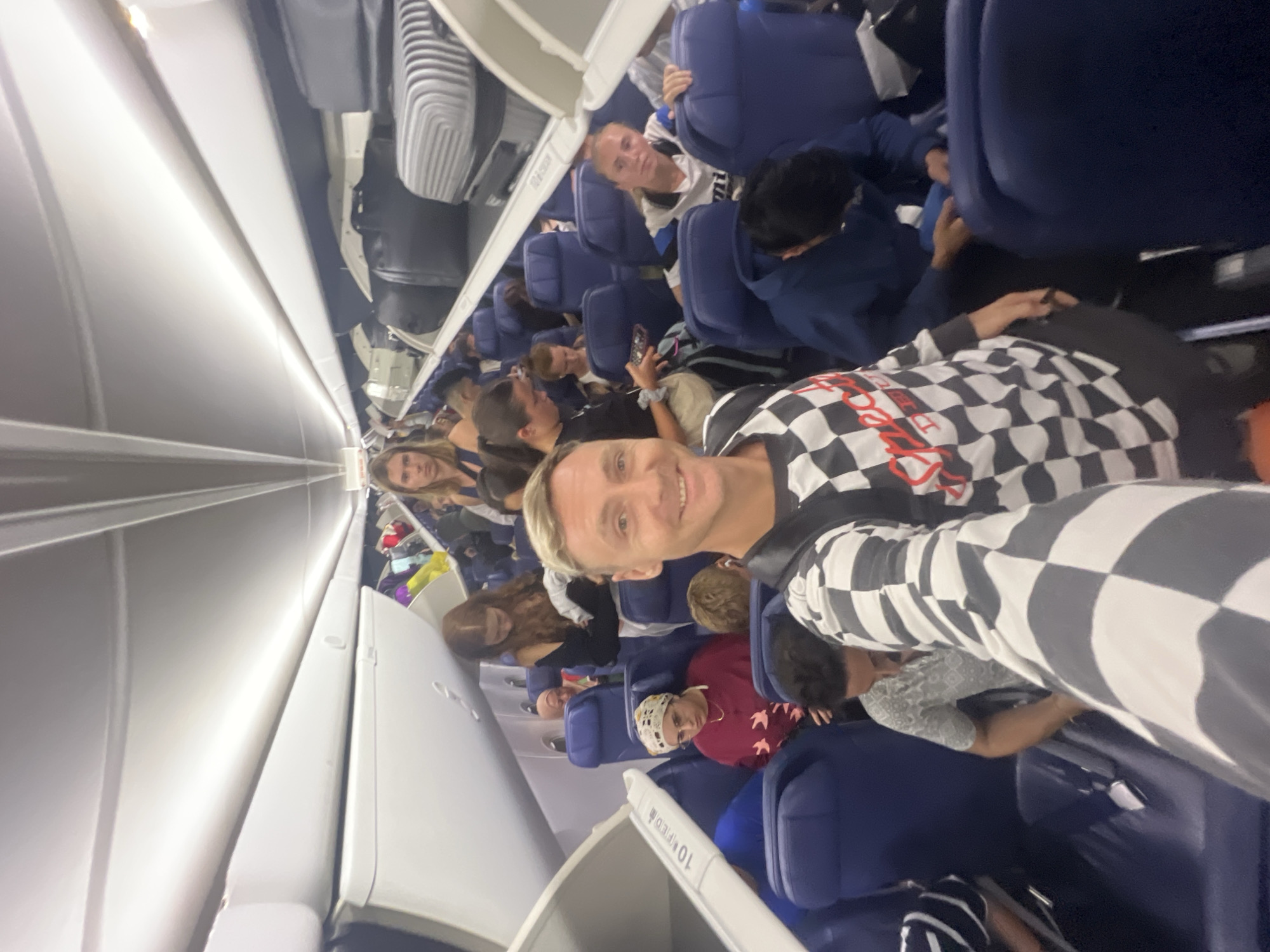 The image captures the interior of a commercial airplane cabin, viewed from a slightly low angle looking down an aisle towards the rear. The scene appears to be taken during boarding, disembarking, or a wait before departure/after arrival, with many passengers visible.
In the immediate foreground, a fair-skinned man with short blonde hair and a wide smile looks directly at the camera. He is wearing a distinctive black and white checkered long-sleeved shirt with some red lettering, partially visible, across the chest (possibly "DREAMER" or similar). He seems to be taking a selfie or posing for a close-up photo.
Behind and around him, numerous other passengers occupy the blue-upholstered seats on both sides of the aisle. Some are looking forward, others are engaged in conversation, or looking towards the camera. To the man's right, a person in a dark blue hoodie is visible. Further down the aisle, a woman with long brown hair is visible on the left, and another person with lighter hair is looking forward from their seat. A person wearing a red top and a light-colored patterned head covering is also seated in the mid-ground.
The overhead compartments, made of light-colored material, are closed, with some luggage visible inside or stored above. One compartment door clearly displays the text "10L." The cabin lighting is artificial, suggesting it could be any time of day, or simply a well-lit interior. The overall atmosphere seems casual, with many individuals and groups present. The location context suggests this flight is in or around Kuta, Indonesia.