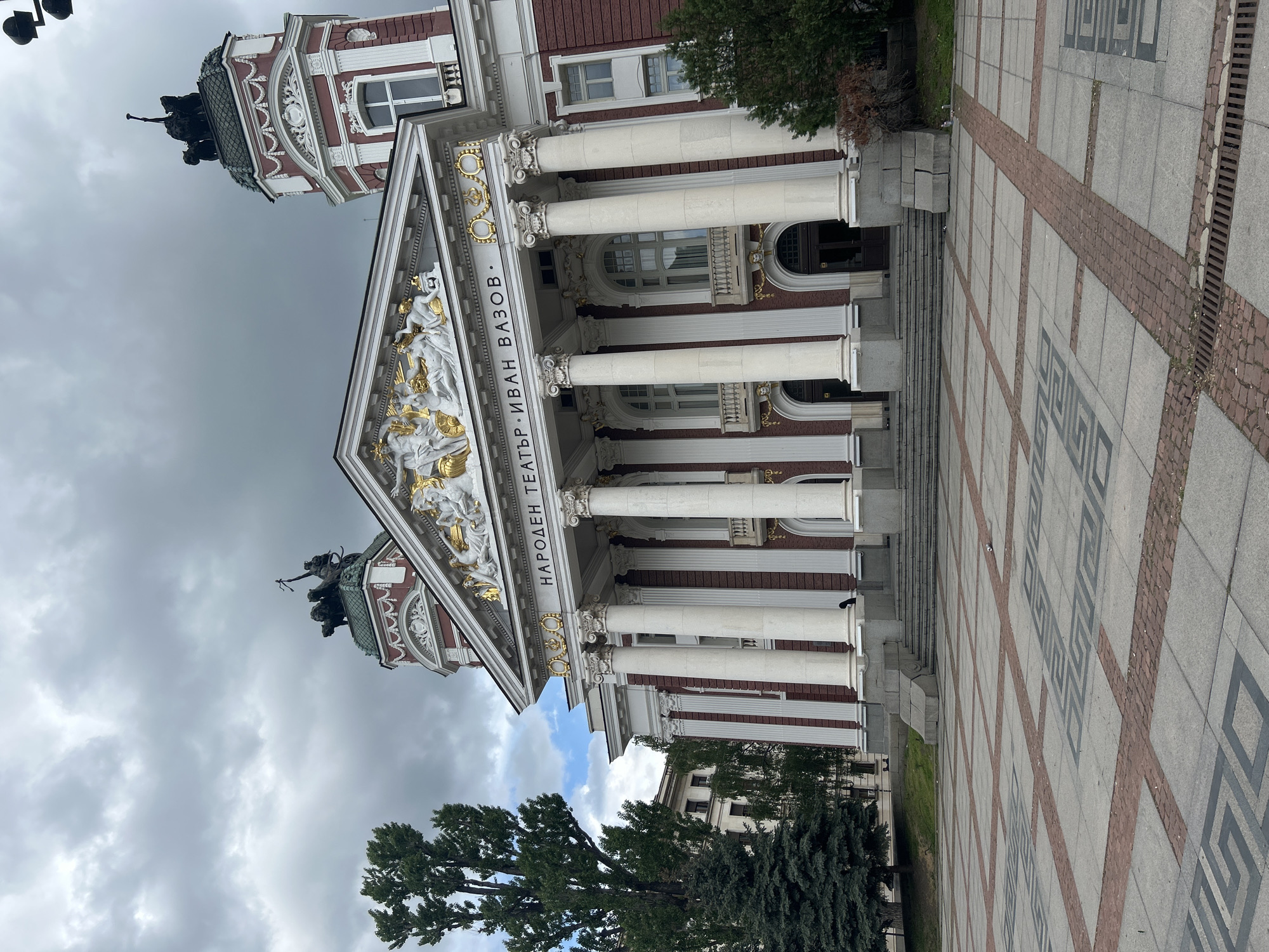 The image captures the Ivan Vazov National Theatre in Sofia, Bulgaria. The building exhibits neoclassical architecture with white columns, ornate gold detailing, and brick-red accents. Statues adorn the roofline. The surrounding area is paved with patterned stones and includes greenery from trees and bushes. The sky is overcast, suggesting a cloudy day.