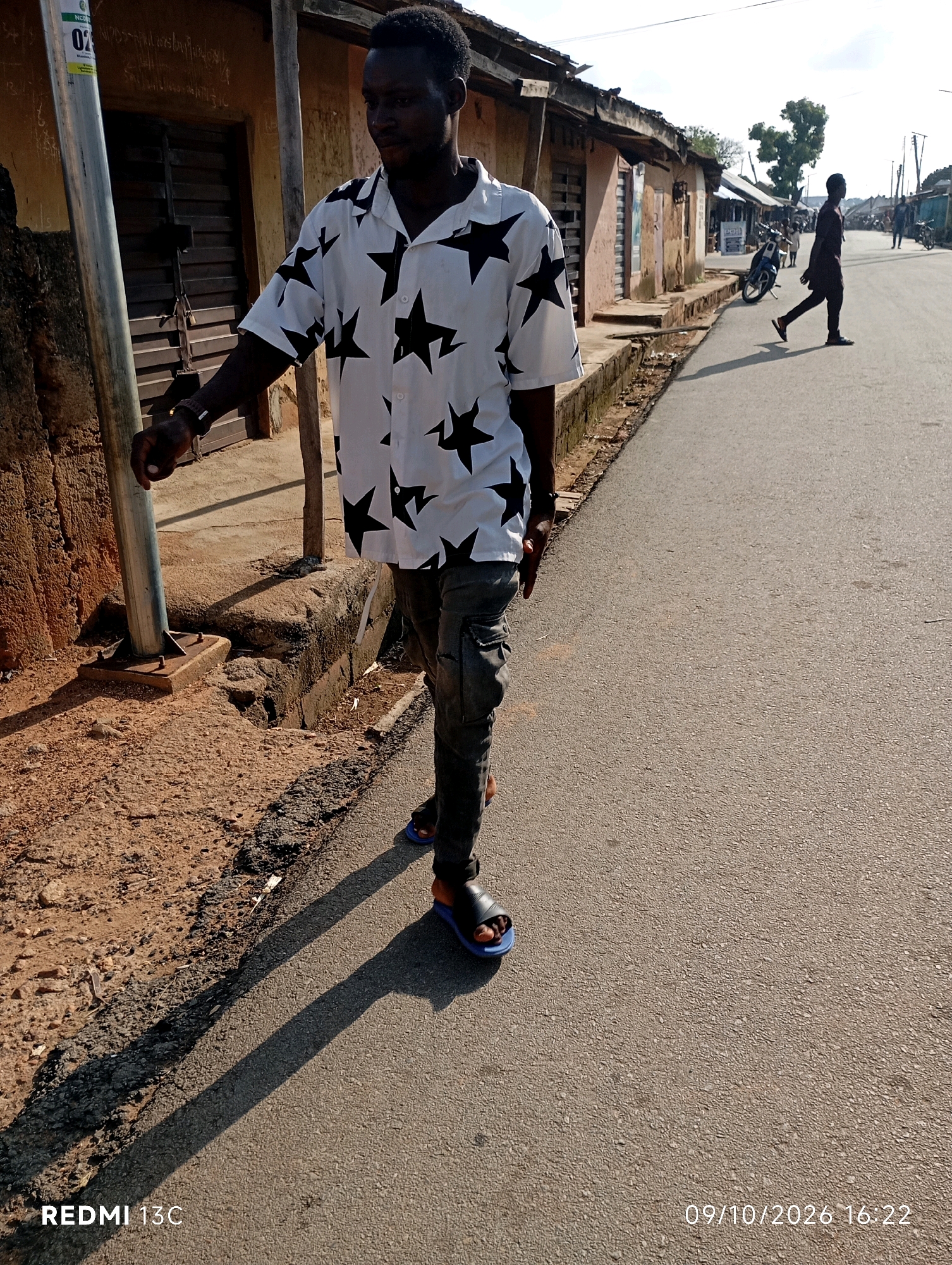 The image captures a street scene in Shendam, Nigeria, under bright daylight. In the foreground, a man wearing a white short-sleeved shirt with black star patterns, dark cargo pants, and black sandals with blue soles is walking on an asphalt road. He is casting a prominent shadow on the road. To his left, there's a dirt path beside a low-lying concrete structure, with a metal pole on the left edge of the frame. The background shows a street lined with single-story buildings with corrugated iron roofs, some appearing weathered. A motorcycle is parked in front of one of the buildings. Further down the street, other people and motorcycles are visible, suggesting a moderately active urban environment. The sky is clear and bright. The timestamp at the bottom right corner of the image reads "09/10/2026 16:22".