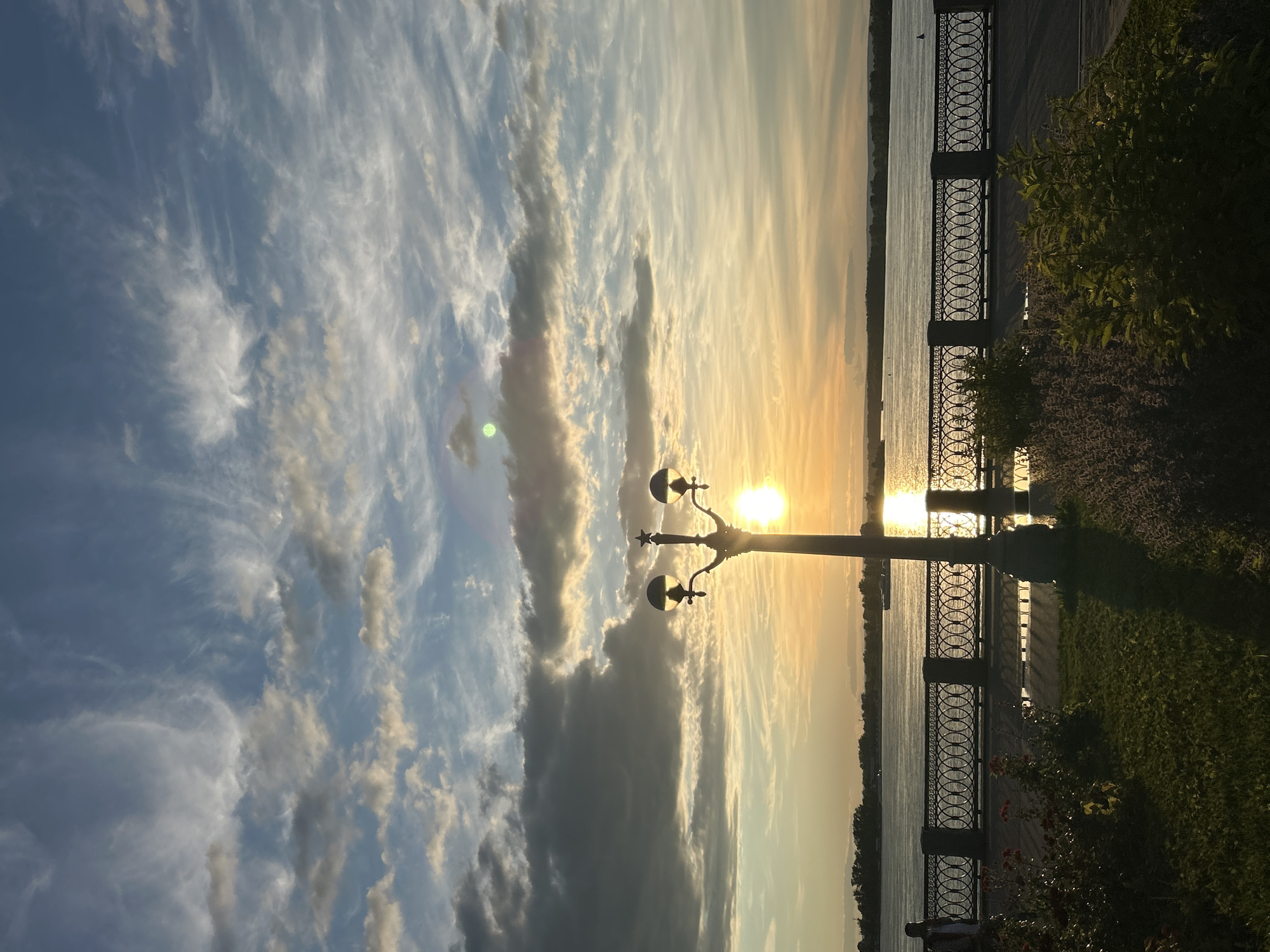 This is a vertically oriented, eye-level, medium shot of a sunset over a body of water. The sun is low in the sky, casting a warm, golden light across the scene. Wispy clouds are scattered across a blue sky, some catching the sunlight and appearing to glow. In the foreground, a lamppost with two globes stands beside a decorative metal fence that runs along a waterfront promenade. The promenade is paved with cobblestones. To the right of the fence, there is a grassy area with some bushes and flowers. The water appears calm, with a slight ripple reflecting the sunlight. A distant treeline is visible on the opposite bank of the water. The overall mood is peaceful and serene, suggesting a tranquil evening by the water. The time of day is sunset, and the weather appears clear. The location cues point to Novokuybyshevsk, Russia. There is no visible text in the image.