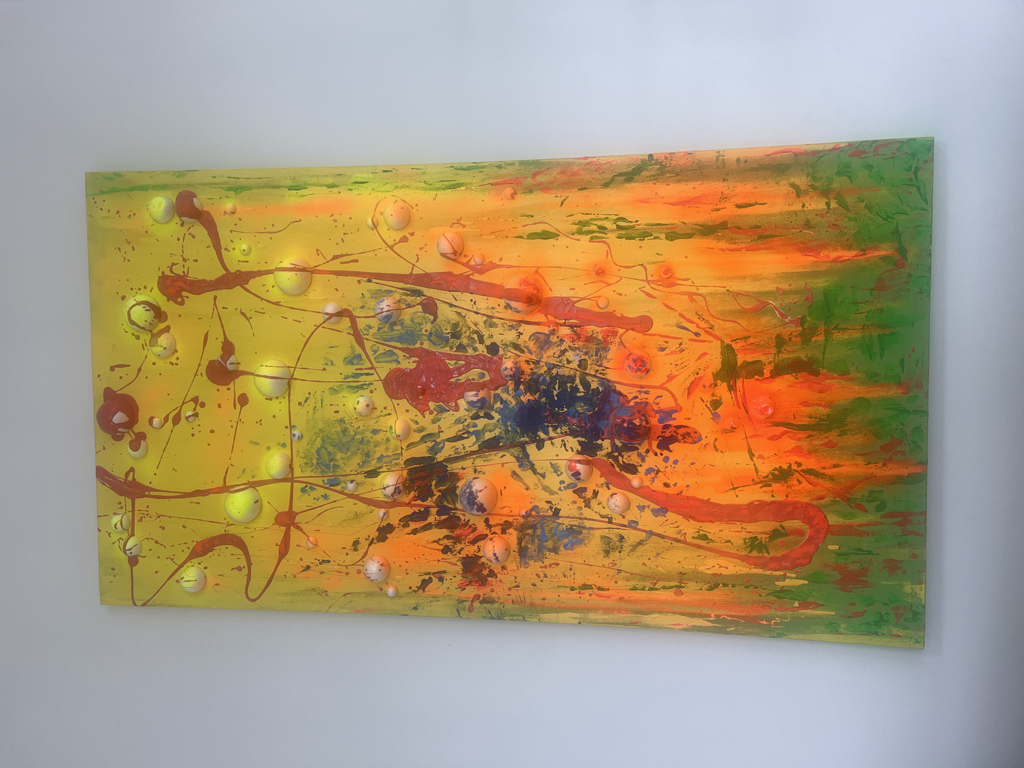 The image shows a rectangular abstract painting hanging on a white wall. The painting features a vibrant mix of yellow, orange, and green hues. The paint application includes broad brushstrokes and splatters. There are round, raised elements of various sizes that appear to be embedded in the painted surface. The painting also has streaks and drips of dark red, blue and brown paint adding depth and dimension. There is no discernible information to place this image in the city of Chaikovsky, Russia.