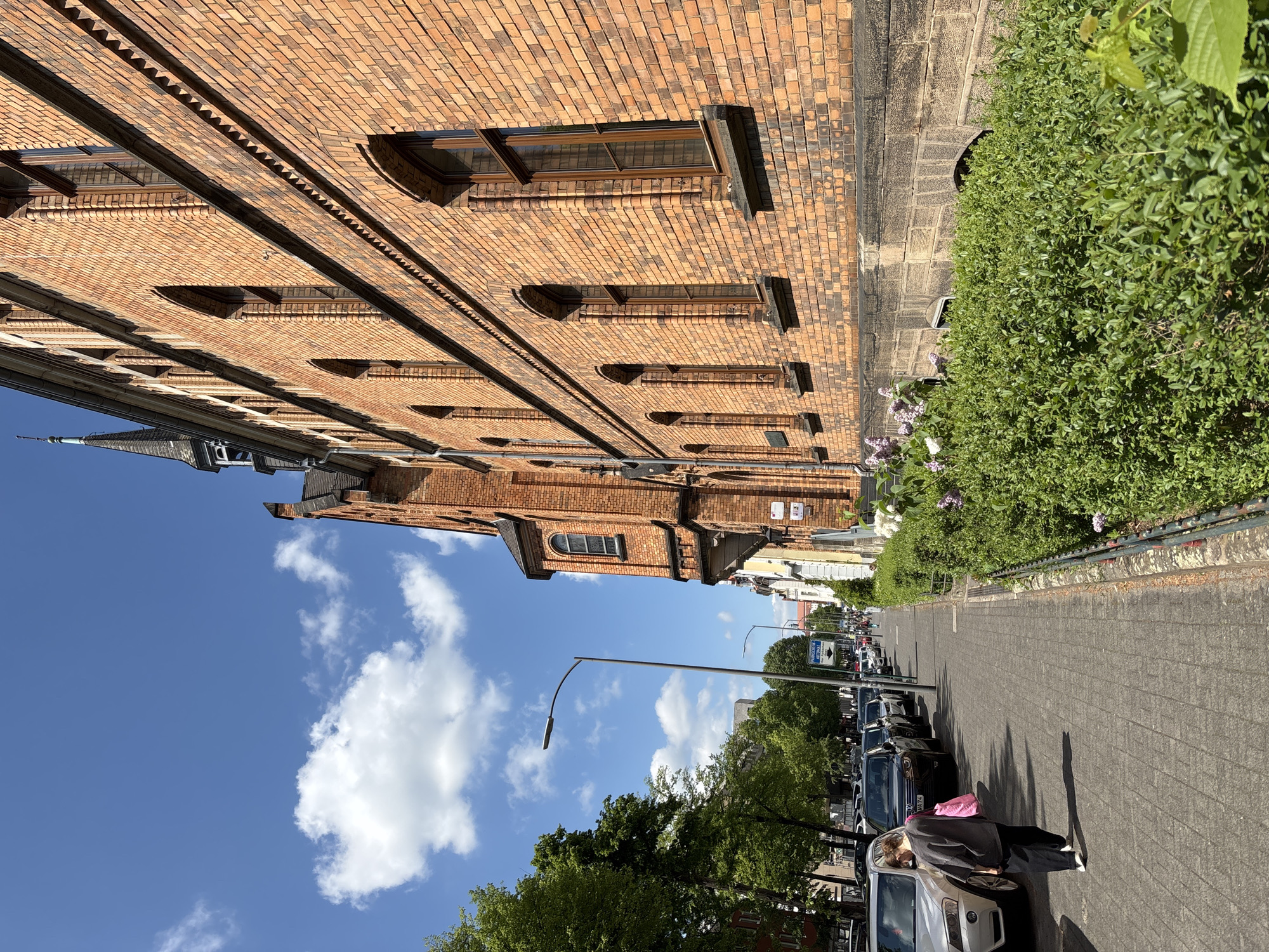 The image depicts a street in Fulda, Germany, lined with parked cars and a brick building with multiple windows. The sidewalk is paved with rectangular stones, and a hedge runs along the side of the building. A person wearing a grey shirt, black pants and carrying a pink bag is standing next to a parked car. The sky is blue with scattered white clouds. A street lamp is visible along the road.
