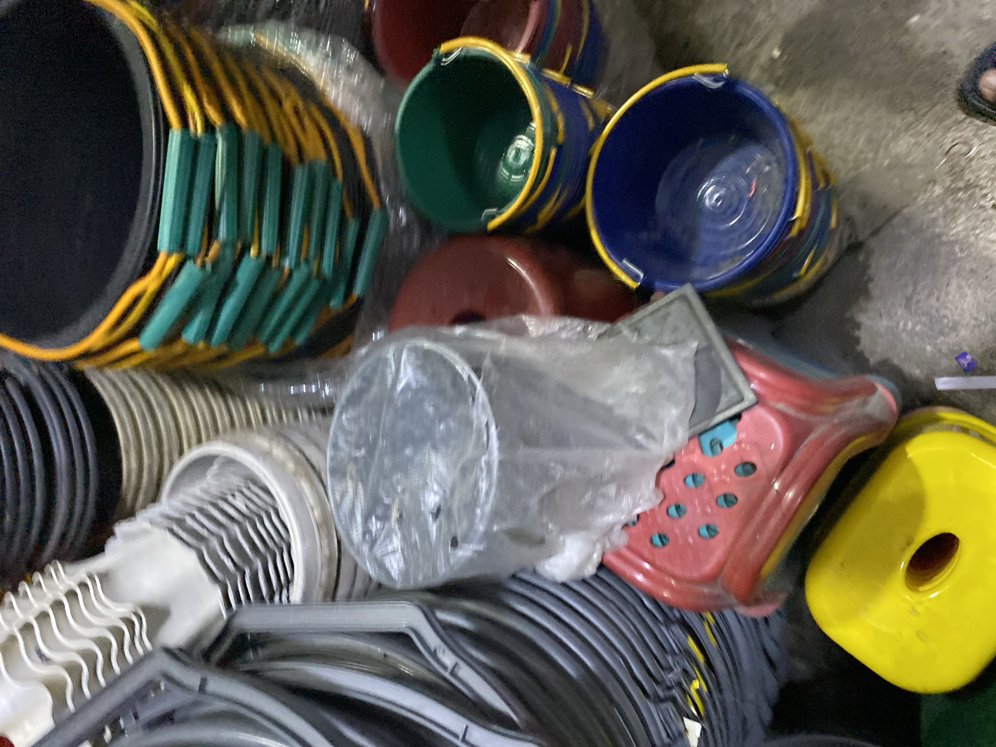 The image depicts an outdoor scene, likely a marketplace or storage area, in Ajuen, Indonesia. The ground is concrete and appears wet in places. The central focus is a collection of various plastic buckets and containers. In the foreground, a bright yellow lid-like object with a central hole is visible. To its left and slightly behind it is a red plastic container with a perforated pattern. To the right and behind the red container are several buckets, including a blue and yellow one, and a green and yellow one. Further back, more buckets are stacked, some with black bases and yellow and green banding. To the left of the image, a stack of white plastic items with a wavy pattern, possibly scoops or bucket handles, are visible, along with grey plastic items that appear to be part of a large container or structure. The overall impression is one of a commercial setting where plastic goods are being stored or sold. No people are clearly visible in the frame.