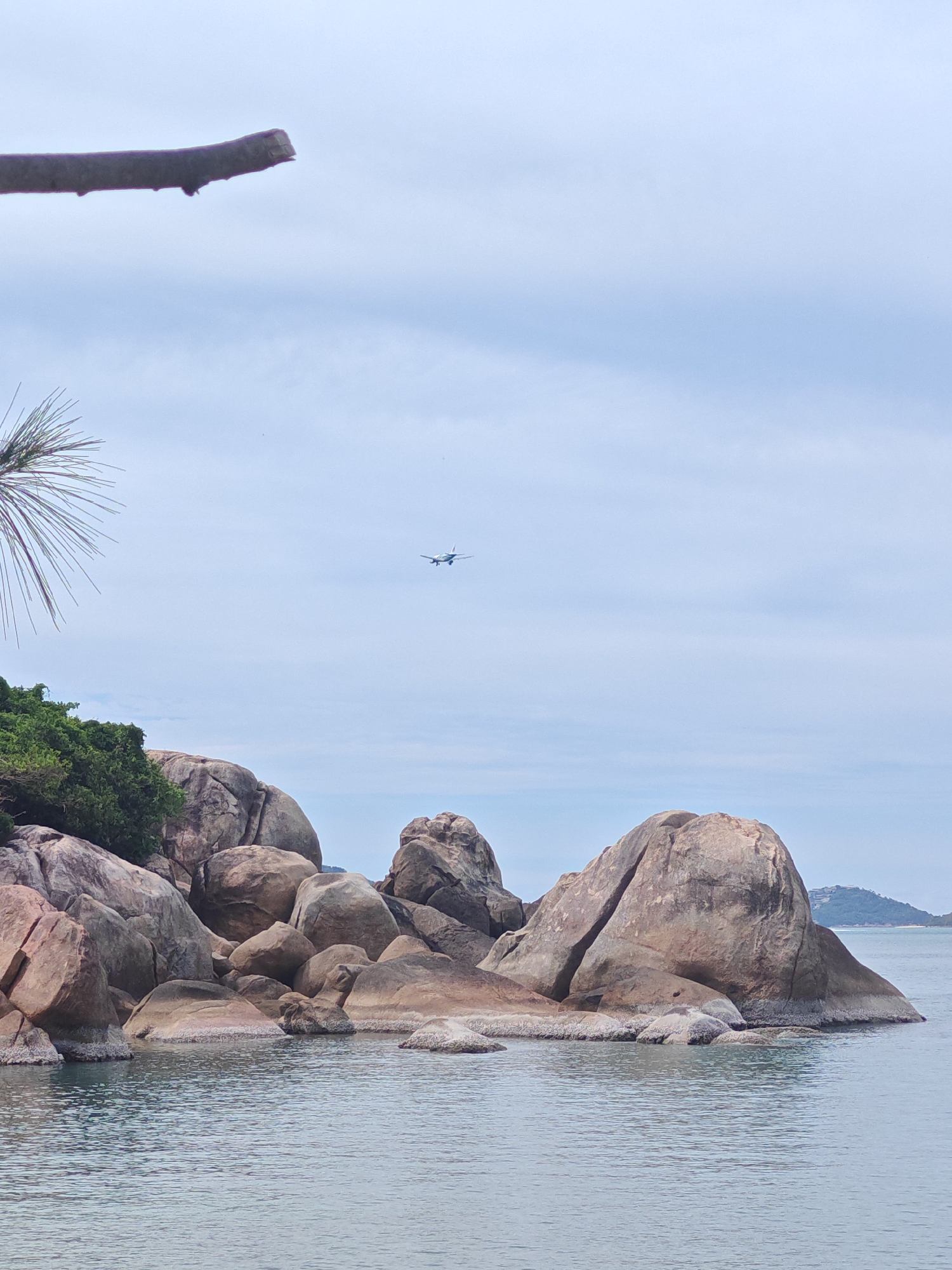 This media file captures a scenic view of a rocky coastline with calm water. The foreground features a cluster of large, weathered boulders of varying sizes, some submerged at their bases. Lush green vegetation covers the top of a rocky outcrop on the left side of the frame.
In the sky, a commercial airplane is visible, appearing small against the expanse of pale blue sky with light cloud cover. A thin, dark tree branch extends from the top left corner of the image, partially obscuring the sky. The water is a tranquil, grayish-blue, reflecting the overcast sky. In the distance, across the water, the faint outline of a landmass with some trees and structures can be seen.
The lighting suggests it is daytime, likely morning or afternoon, with diffused light due to the cloudy conditions. The overall mood is serene and natural. The location is identified as Baan Chaweng Noi, Thailand, which is consistent with the tropical rocky coastline and the presence of lush vegetation. No text is visible in the image.