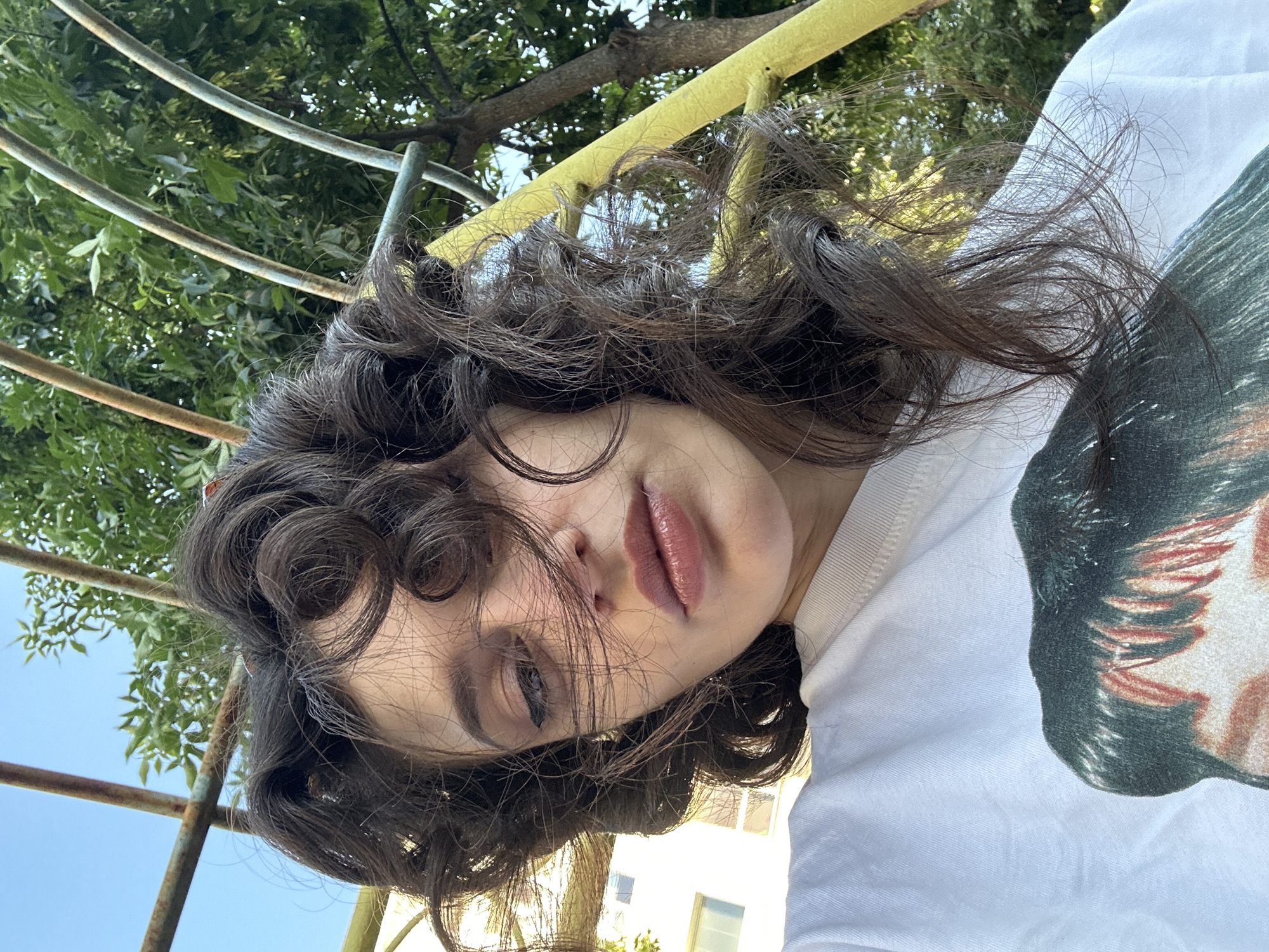 A young woman with dark, curly hair and winged eyeliner takes a selfie outdoors. She is wearing a white t-shirt with a graphic print of a person's face. She appears to be sitting on or near a yellow metal structure, possibly part of a playground or outdoor seating. In the background, there are green trees and a clear blue sky, with a building visible to the left. The scene suggests a casual, daytime setting, potentially in a park or residential area.