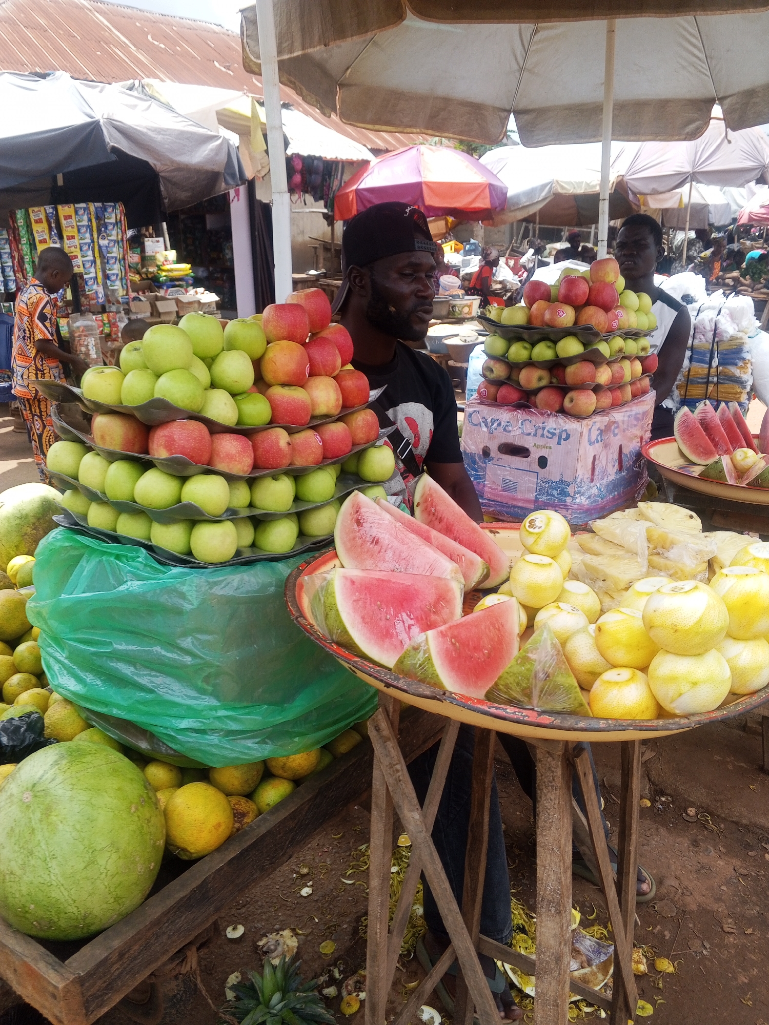 The image depicts a busy outdoor market stall in Kabba, Nigeria, with a focus on fresh fruit. A man wearing a black baseball cap and a black t-shirt stands behind a display of apples and other fruits. He is looking slightly to his left. In front of him, a tiered stand is filled with apples, a mix of red and green. Below the apples, a large green plastic bag holds more produce, partially obscuring a large green watermelon on the left side of the frame. To the right of the apples, there is a tray of sliced watermelon, some citrus fruits, and what appears to be pineapple chunks wrapped in plastic. Another man, partially visible on the far left, is wearing a colorful patterned shirt and is interacting with items on a shelf behind the fruit stall. The market environment is suggested by the presence of various umbrellas, other stalls in the background, and the general atmosphere of commerce. The ground appears to be dirt, littered with discarded fruit peels and debris.