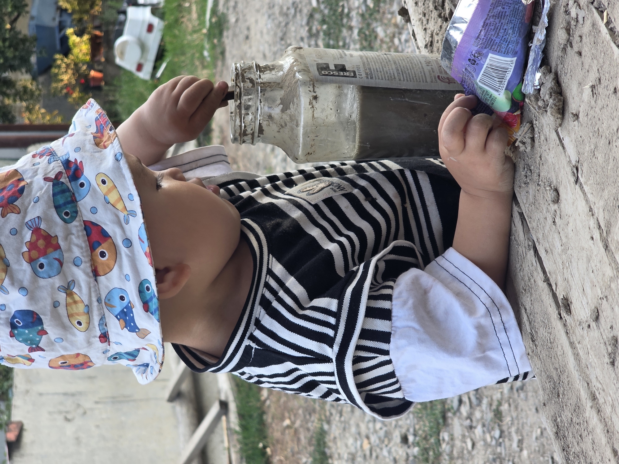 A young child, wearing a white bucket hat with a fish pattern and a black and white striped shirt, is intently engaged in an activity. The child's right hand is inside a glass jar containing a murky liquid and a stick, suggesting they are stirring or playing with the contents. To the right of the jar, a crumpled purple wrapper lies on a wooden surface. The environment appears to be outdoors, with a gravelly ground and hints of greenery in the background, along with a white toy vehicle. The image is taken from a medium close-up perspective, focusing on the child and their immediate surroundings. The overall scene suggests a moment of childhood exploration and play, possibly in a yard or a semi-rural setting.
