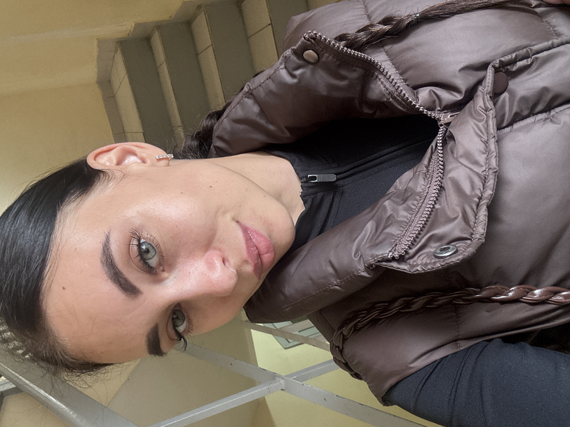 A forensic examination of the image reveals a close-up portrait of a young adult female, approximately 20-35 years old, positioned indoors within what appears to be a residential or public stairwell in the city of Chaikovsky, Russia. The subject has dark hair, styled in a braid visible over her right shoulder, light-colored eyes (appearing blue-green), and well-defined eyebrows. Her skin tone is fair, with a few visible freckles across her nose and cheeks. She wears a small, diamond-studded hoop earring in her visible right earlobe. Her lips are slightly parted, showing a faint pink hue, indicative of lip product. She is attired in a black long-sleeved garment underneath a brown, quilted puffer vest, which features a central zipper closure, snap buttons on the collar, and a decorative brown braided trim running vertically along the right side of the garment. The background consists of light yellow-painted walls and a grey metallic railing system on the left, with concrete or painted steps visible ascending in the upper right quadrant of the frame. The lighting is soft and ambient, illuminating the subject's face directly.