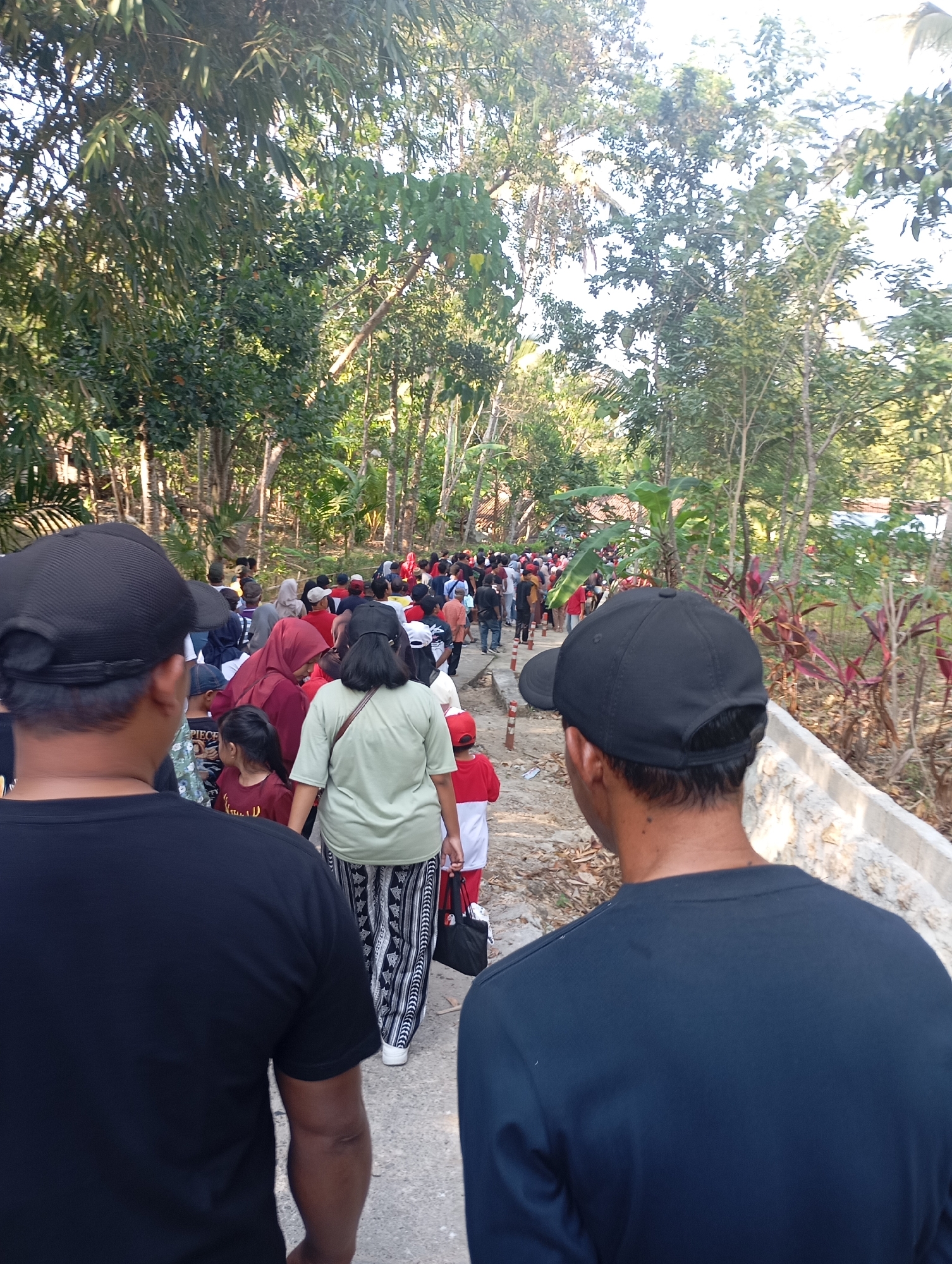 The image depicts a gathering of people walking along a path in a rural or semi-rural area, likely in Ketepung, Indonesia, given the context provided. The scene is characterized by lush vegetation, with trees and various plants lining both sides of the path. The path itself appears to be made of compacted earth or gravel, with a concrete structure on the right side, possibly a low wall or raised walkway.
In the foreground, two individuals are seen from behind, both wearing black caps. The person on the left is wearing a black t-shirt, and the person on the right is wearing a dark blue long-sleeved shirt. They are positioned to observe the group ahead of them.
The main group of people is walking away from the camera, creating a sense of movement and procession. This group consists of a diverse mix of individuals, including adults and children. Several people are wearing brightly colored clothing, such as red and white outfits, and a woman in the middle is wearing a green shirt and patterned pants. Some individuals are carrying bags, suggesting they may be on a journey or attending an event. The overall atmosphere seems to be one of a community gathering or an organized activity. The lighting suggests it is daytime, with sunlight filtering through the trees, creating dappled shadows on the path.