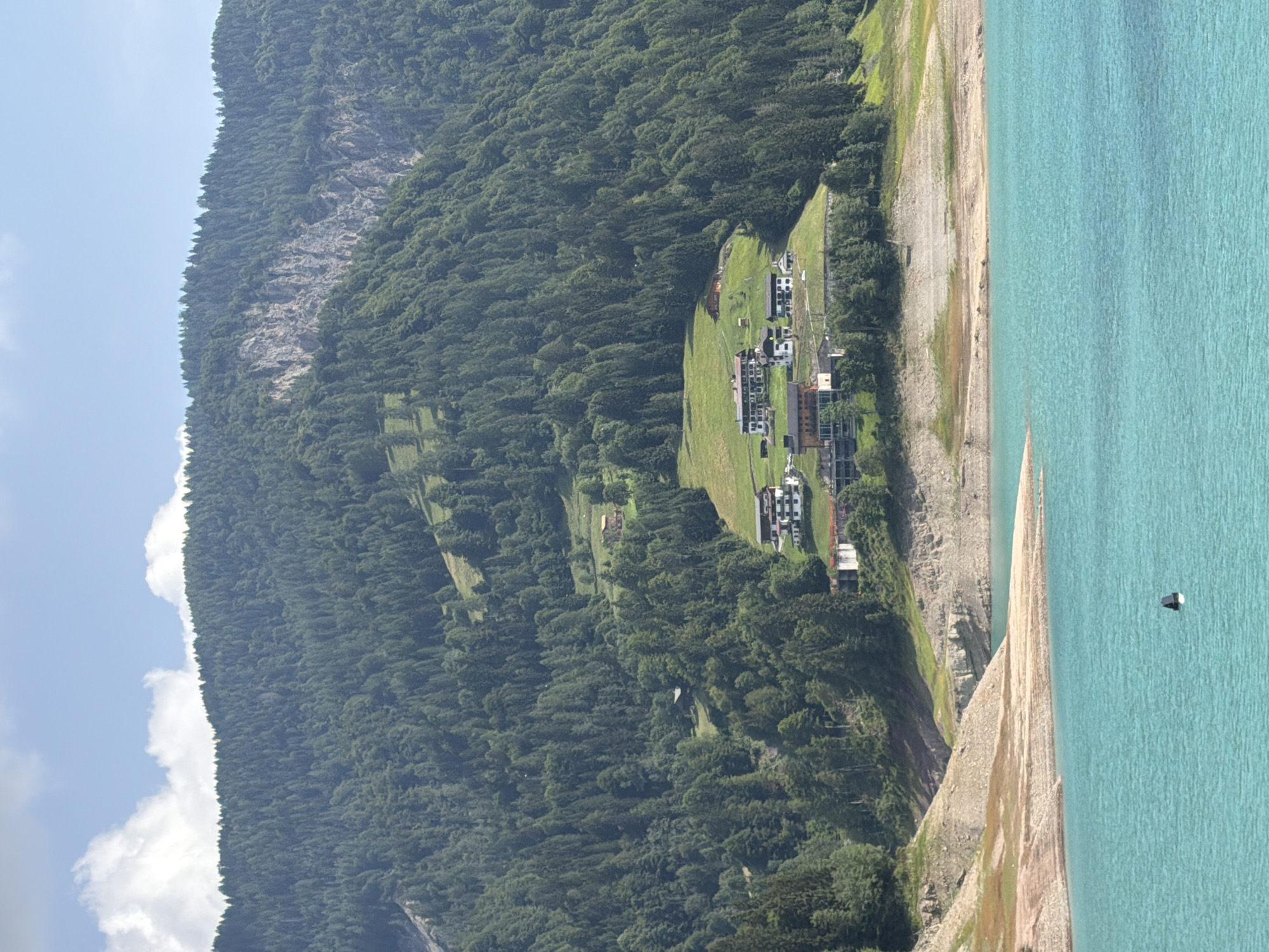 The image shows a vast expanse of turquoise water in the foreground, with a rocky, dry shoreline leading up to a steep, tree-covered mountainside. Several buildings are visible nestled on a grassy slope halfway up the mountain. The mountain range is densely forested with evergreens, and a rocky cliff face is apparent on the upper left side of the mountain. The sky above is partly cloudy with white clouds against a blue background. A black and white buoy is floating in the water. The scene depicts a serene natural landscape with human habitation integrated into the mountainside. The location is identified as Lateis / Latais, Italy.