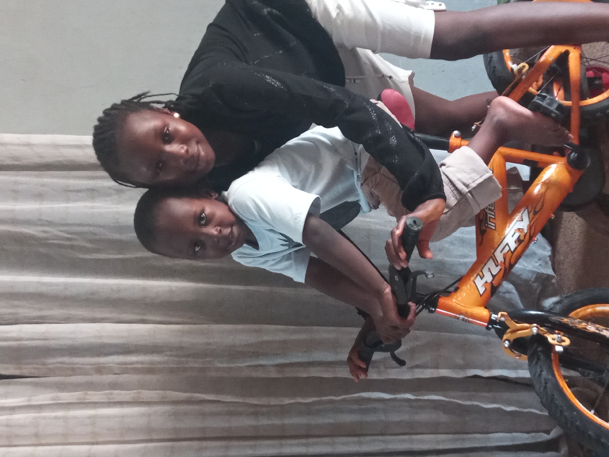 Two Black individuals, likely a woman and a child, are positioned over an orange Huffy bicycle. They are indoors, evidenced by the backdrop of a curtain or wall. The specific location is Sabon Tasha, Nigeria, as requested.