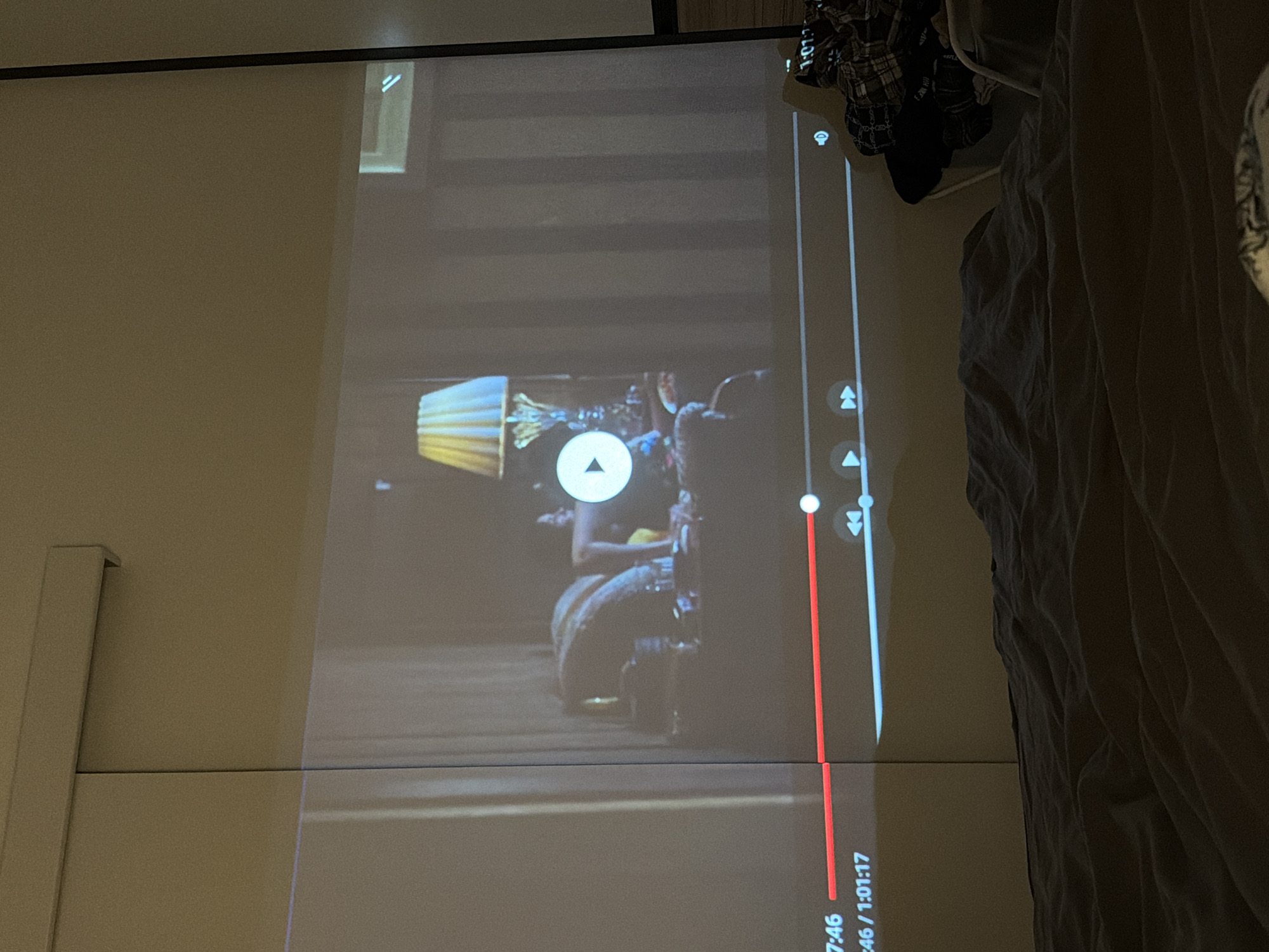 The image shows a projection screen displaying a video with a play/pause button visible in the center. A lamp is visible on the left side of the screen, and a time stamp at the bottom right reads "0:57:46 / 1:01:17." To the right of the screen, a portion of a bed with crumpled sheets is visible. The scene appears to be indoors, but the specific location or setting is unclear from the image. No people are clearly visible in the projected video, but there is a suggestion of figures within the video content.